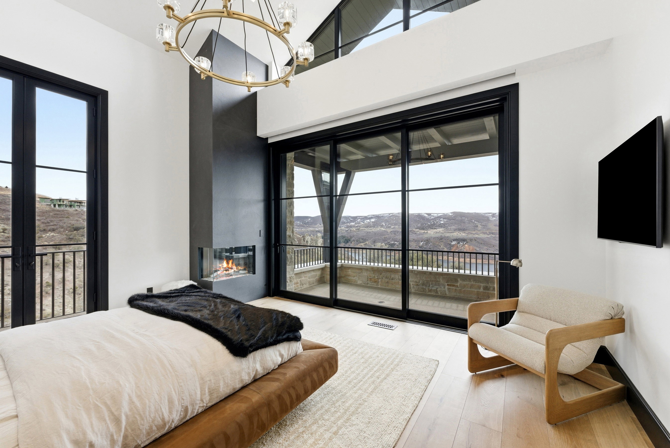 Spacious room featuring a contemporary fireplace with a glass enclosure, a large sliding glass door opening to a private balcony, a separate exterior door with a balcony, light wood-finish flooring, and an overhead chandelier with clear glass globes