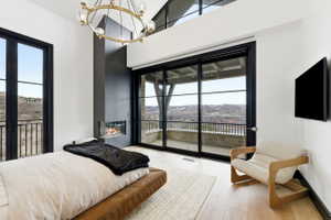 Spacious room featuring a contemporary fireplace with a glass enclosure, a large sliding glass door opening to a private balcony, a separate exterior door with a balcony, light wood-finish flooring, and an overhead chandelier with clear glass globes