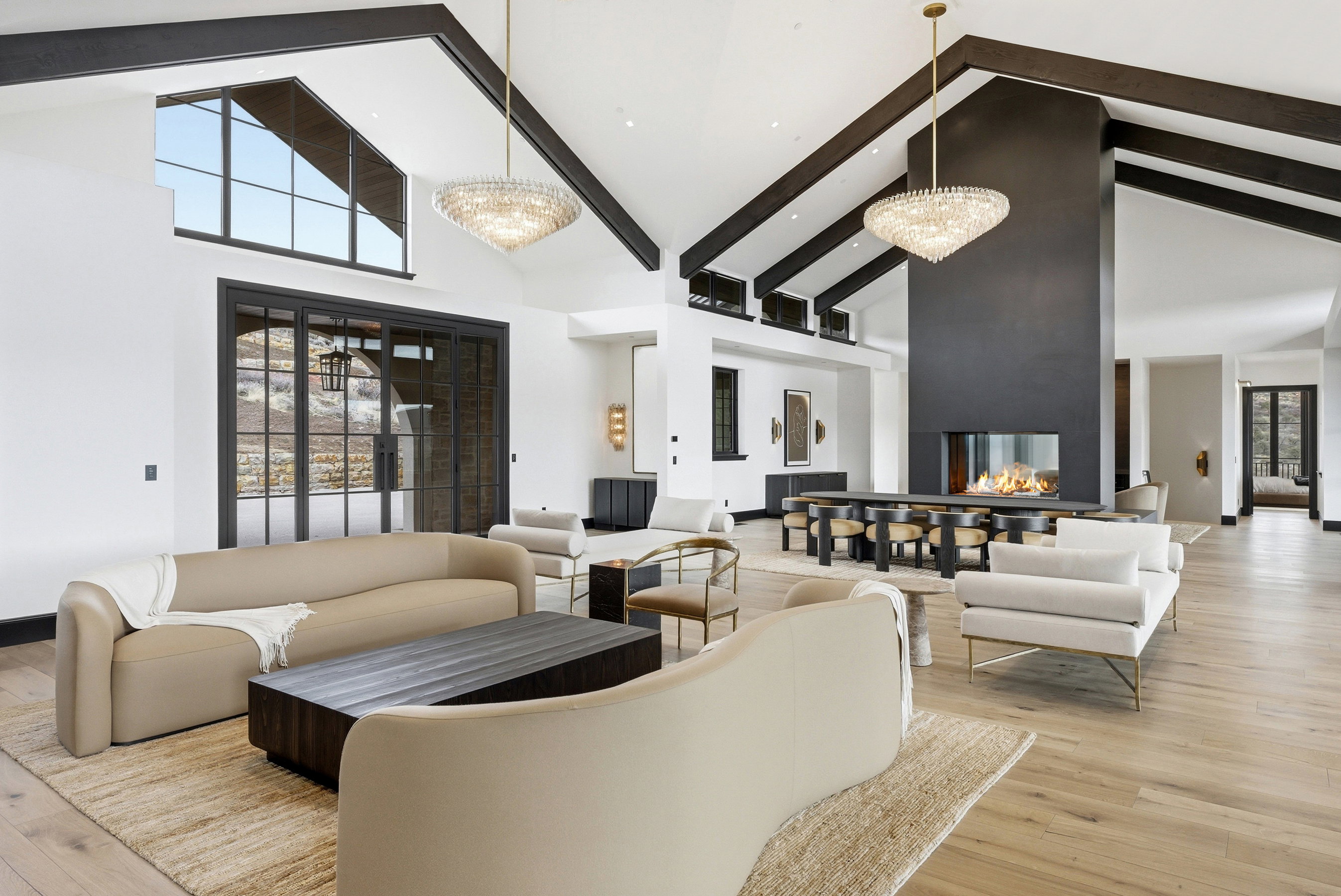 Vaulted great room with exposed dark wood beams, light wood-finish flooring, two tiered crystal chandeliers, a central fireplace with a horizontal glass-enclosed firebox, and a dining table with seating for eight