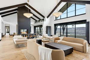 Spacious living area featuring a vaulted ceiling with exposed wood beams, a prominent fireplace with a dark finish, light wood-finish flooring, large multi-pane windows, and elegant chandeliers