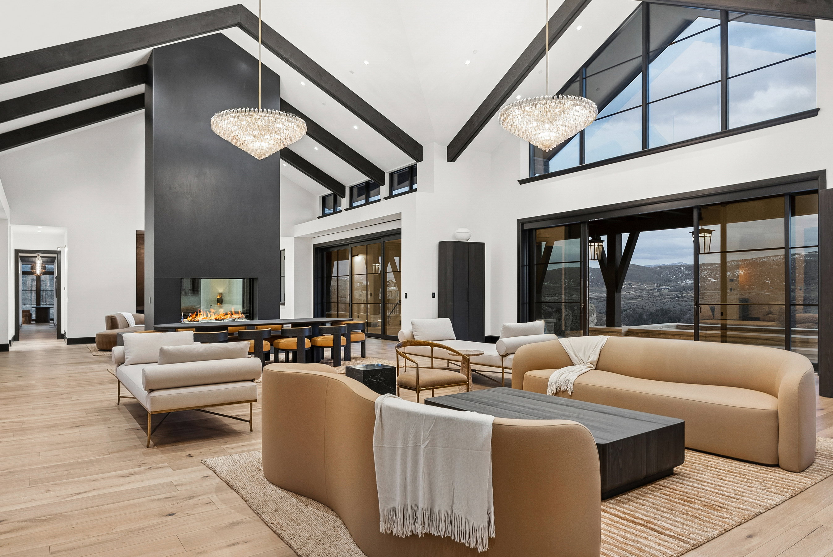 Spacious living area featuring vaulted ceilings with exposed dark wood beams, light wood-finish flooring, and a floor-to-ceiling dark matte fireplace surround