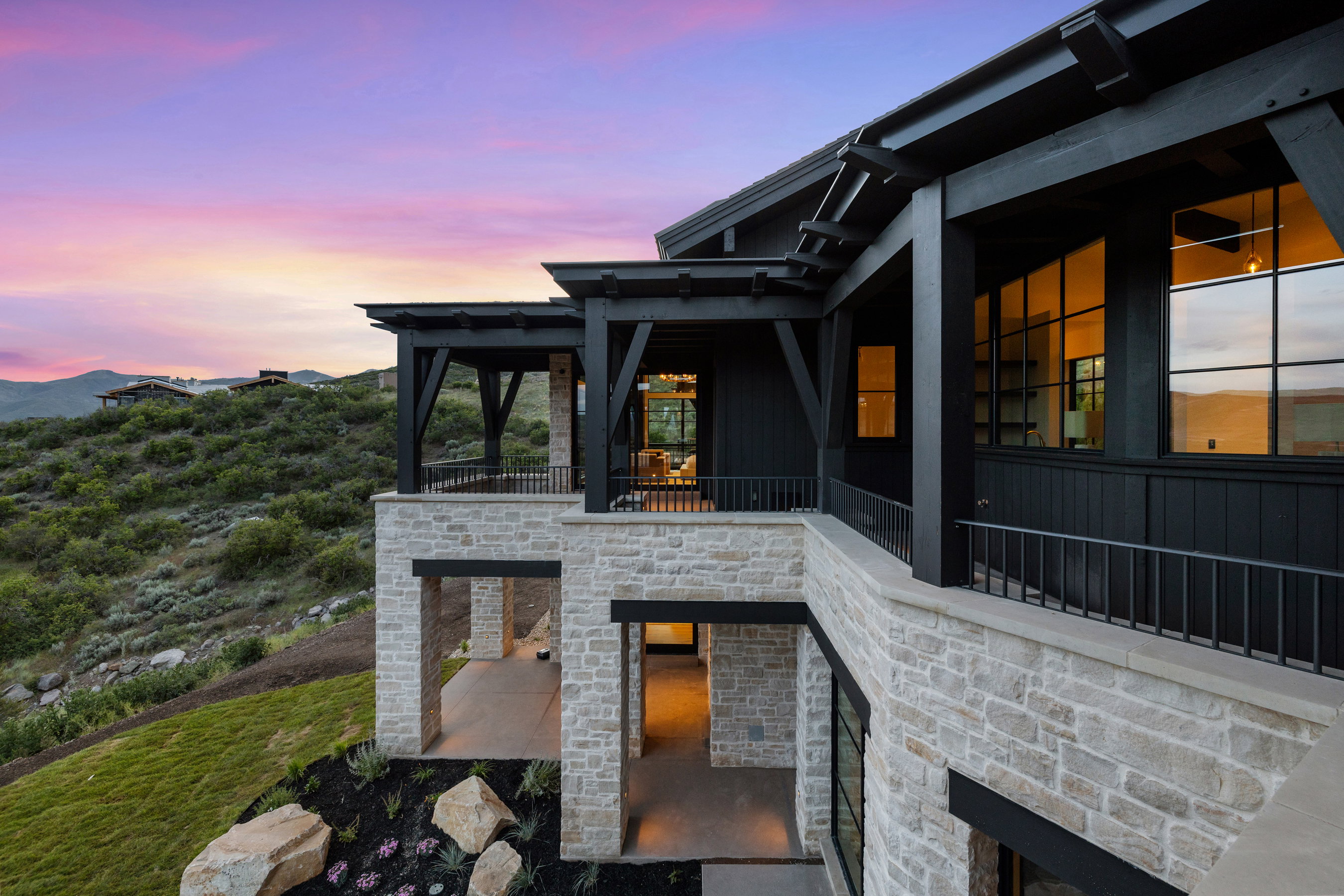 Multi-level exterior featuring natural stone veneer, black wood siding, extensive grid-pattern windows, and covered patios with metal railings