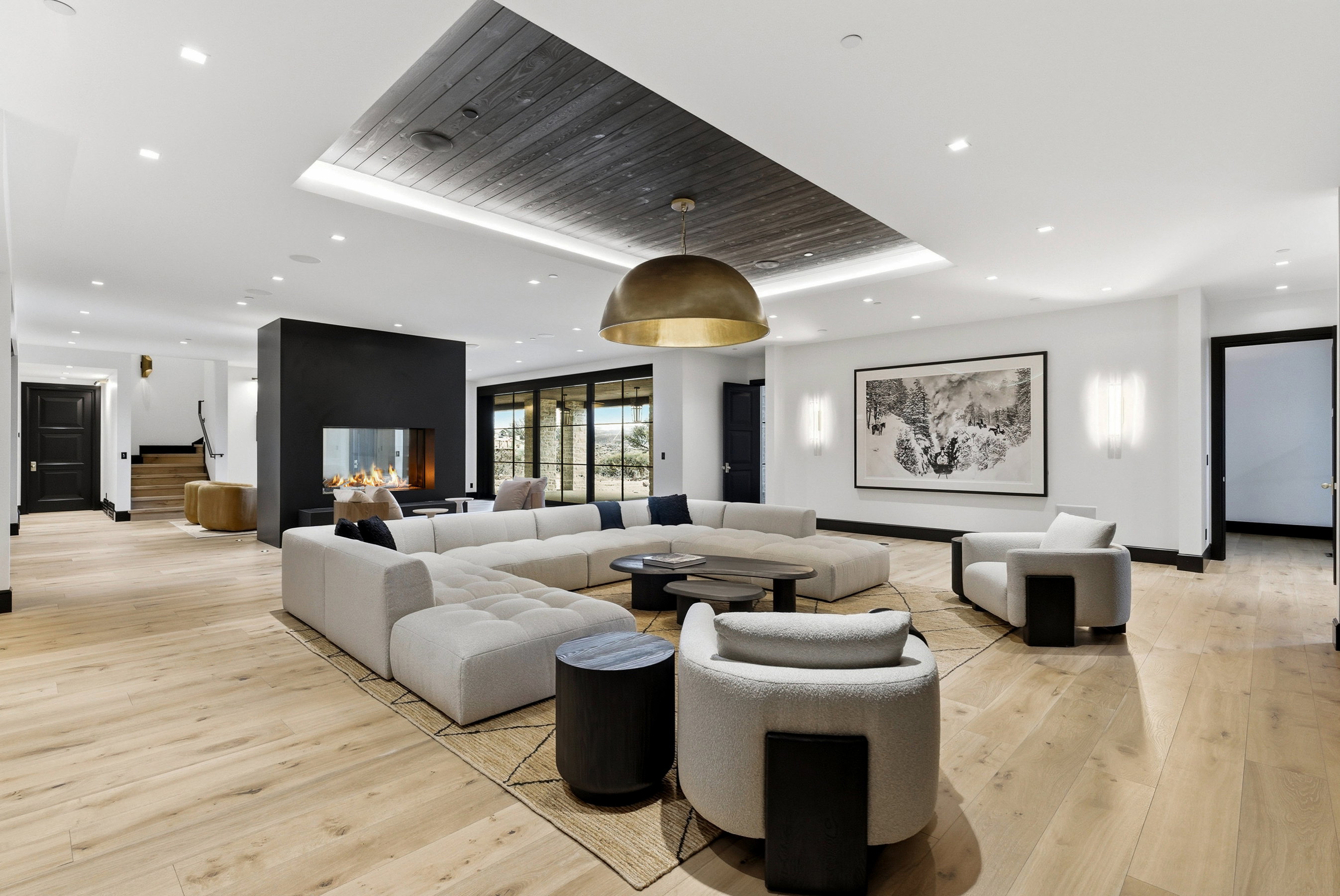 Spacious living area featuring light wood-finish flooring, a recessed ceiling with dark wood-paneling, a large brass dome pendant light, a modern fireplace with a dark surround, and wall-mounted light fixtures