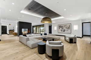 Spacious living area featuring light wood-finish flooring, a recessed ceiling with dark wood-paneling, a large brass dome pendant light, a modern fireplace with a dark surround, and wall-mounted light fixtures