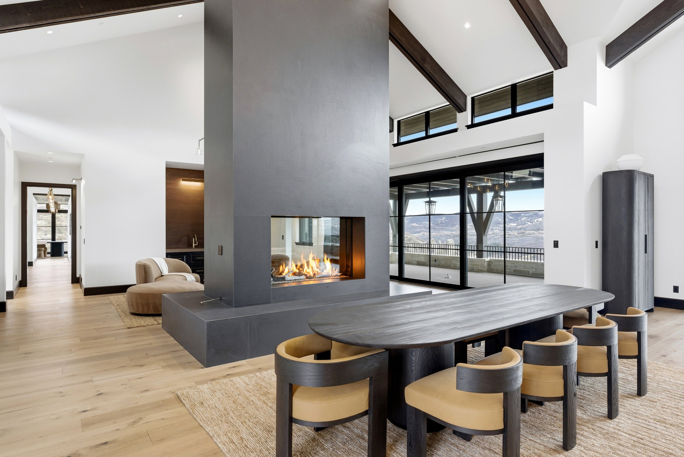 Expansive living area featuring a central four-sided fireplace, wood-finish flooring, exposed ceiling beams, and a black-framed sliding glass door leading to an outdoor space