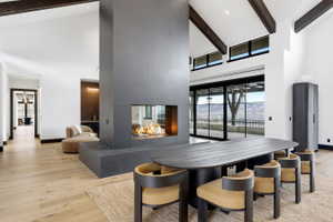 Expansive living area featuring a central four-sided fireplace, wood-finish flooring, exposed ceiling beams, and a black-framed sliding glass door leading to an outdoor space
