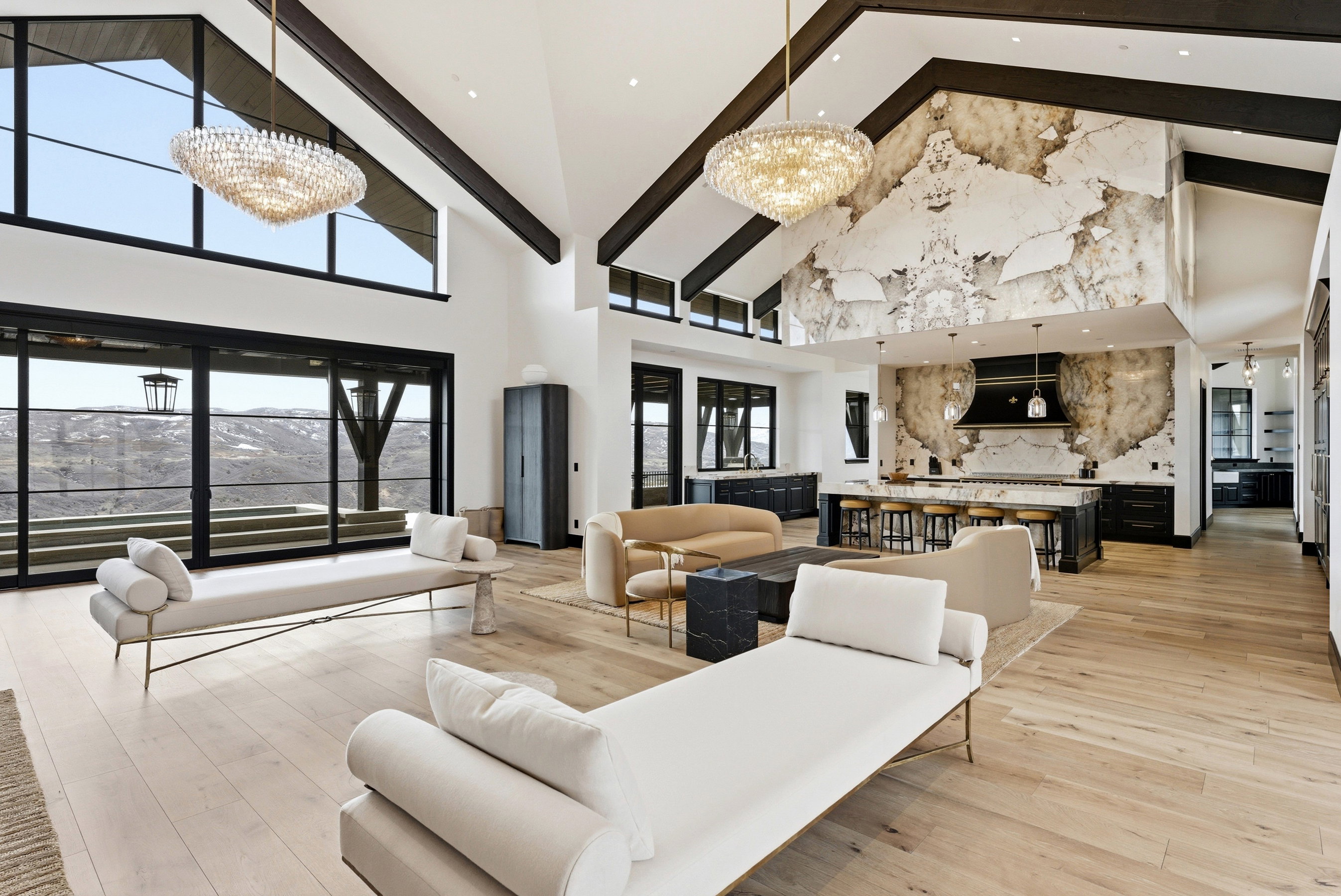 Vaulted ceilings with exposed dark wood beams, expansive floor-to-ceiling windows, light wood-finish flooring, a kitchen island with a stone countertop, and a striking stone accent wall