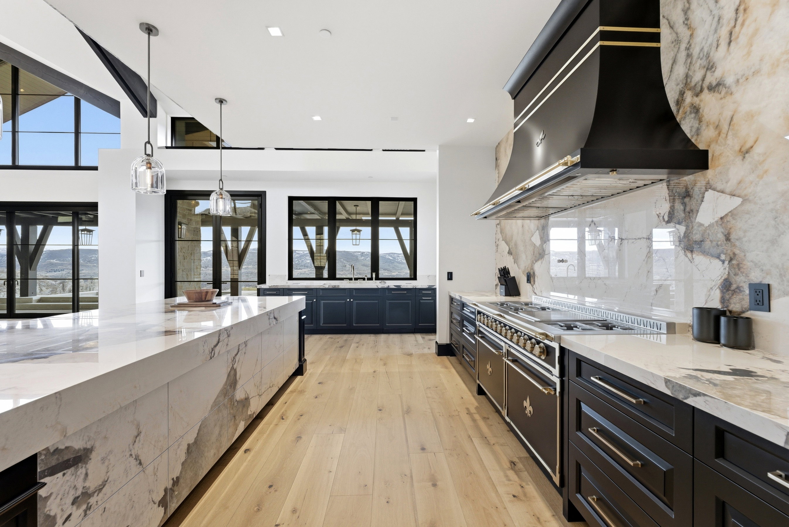 Gourmet kitchen featuring a substantial central island with waterfall-edge countertop, light wood-finish flooring, custom black cabinetry, a professional-grade range, and a coordinating range hood with gold-tone accents