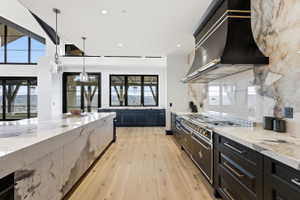 Gourmet kitchen featuring a substantial central island with waterfall-edge countertop, light wood-finish flooring, custom black cabinetry, a professional-grade range, and a coordinating range hood with gold-tone accents