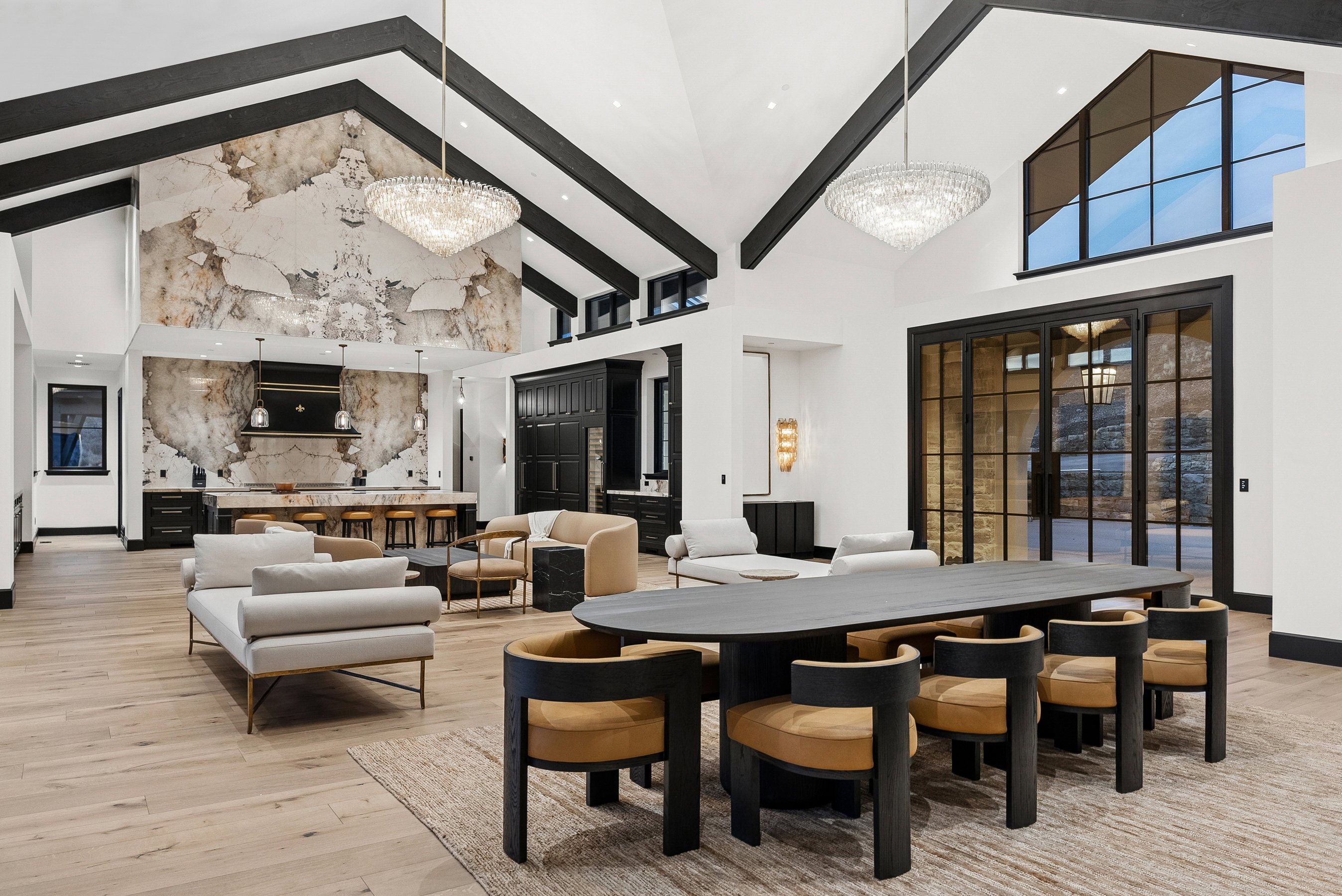 Spacious open-concept interior featuring light wood-finish flooring, vaulted ceilings with exposed black beams, and large-format black-framed windows