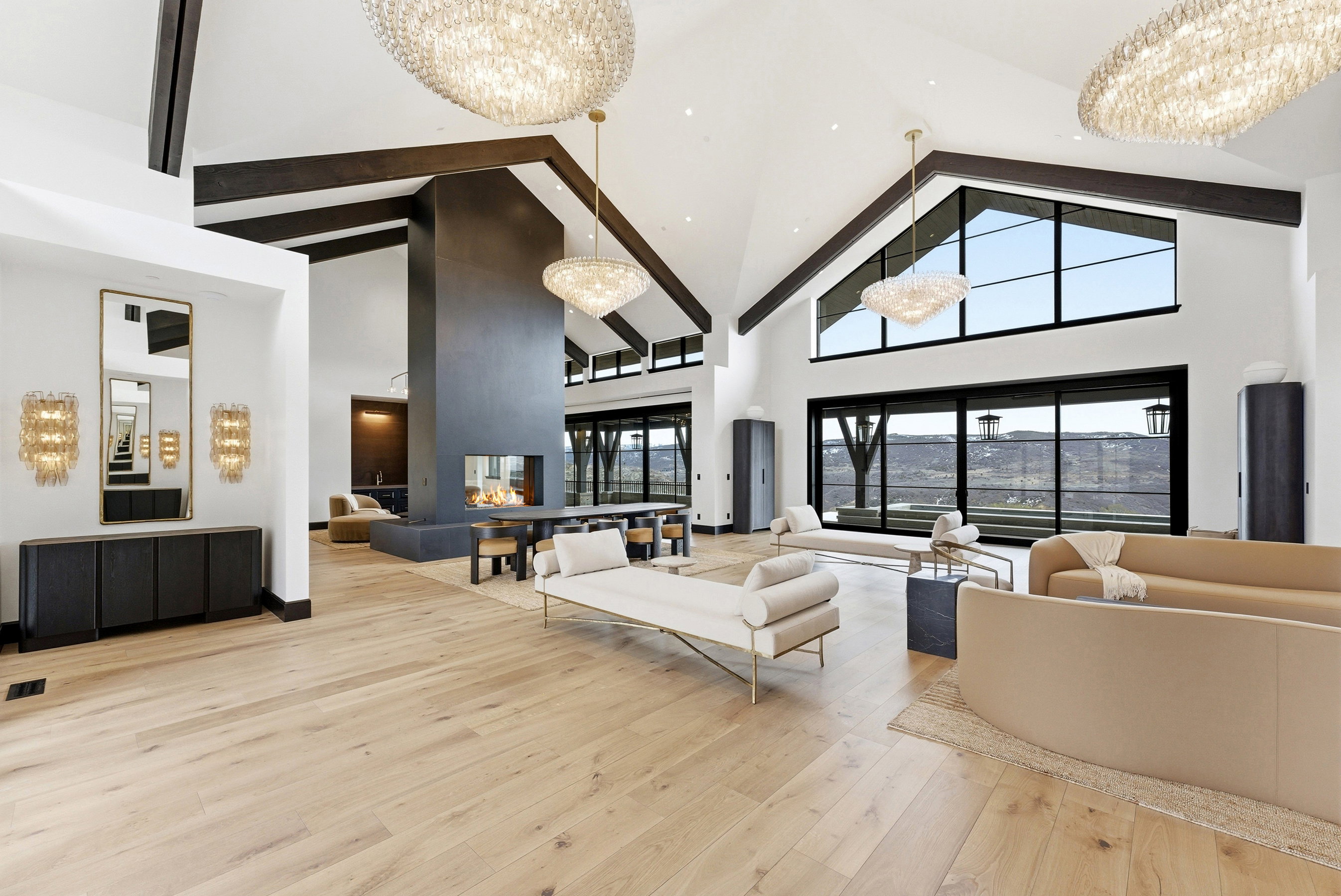 Expansive living space featuring light wood-finish flooring, a vaulted ceiling with dark wood beams, and a floor-to-ceiling fireplace with an integrated hearth