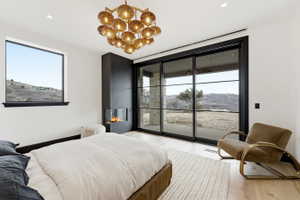 Contemporary bedroom featuring a linear electric fireplace, expansive sliding glass doors, a large picture window, a sophisticated multi-bulb chandelier, and light wood-finish flooring