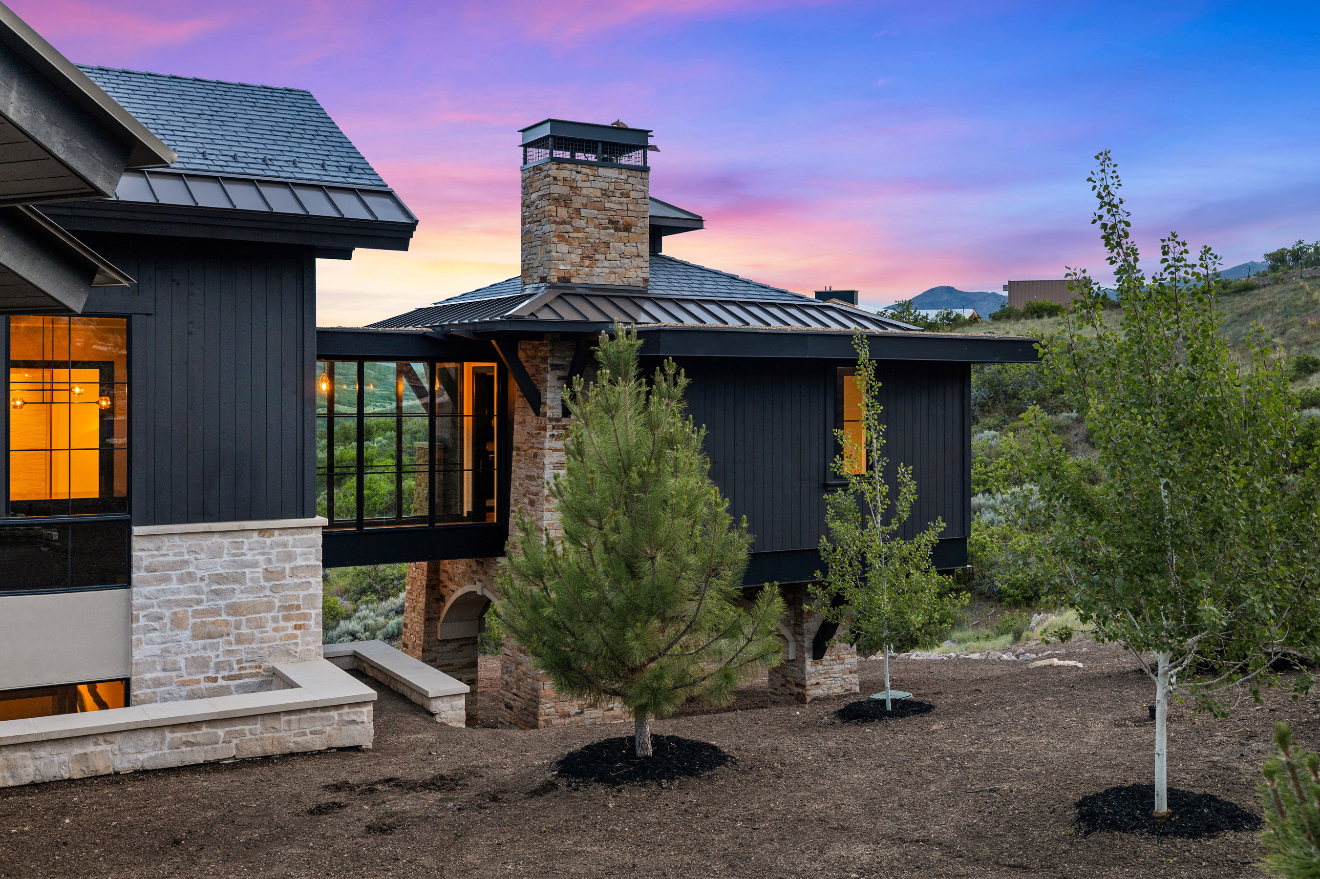 Modern exterior featuring black vertical siding, natural stone accents, a glass-enclosed walkway, and a metal roof