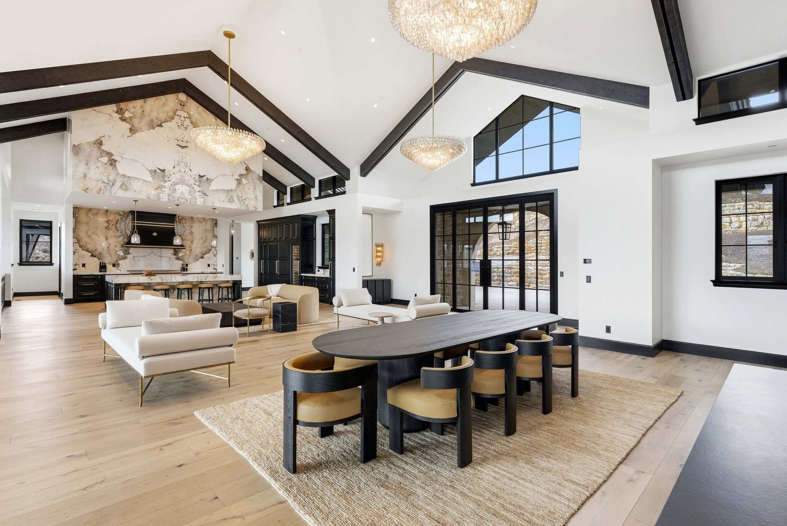 Vaulted ceiling with exposed beams, light wood-finish flooring, full-height decorative stone wall, black framed windows, and a black framed glass door system