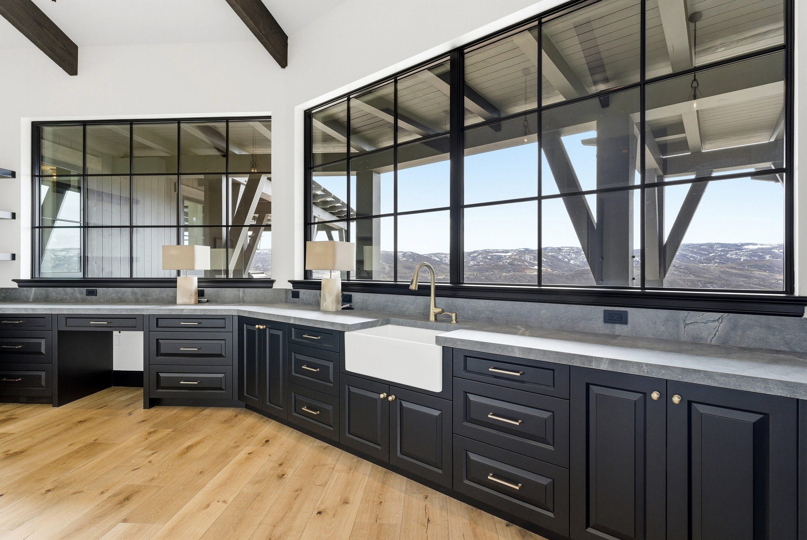 Spacious room featuring multiple large grid-pattern windows, dark cabinetry with gold-tone hardware, a white farmhouse sink with a gold-tone faucet, stone-look countertops, and wide plank wood-finish flooring