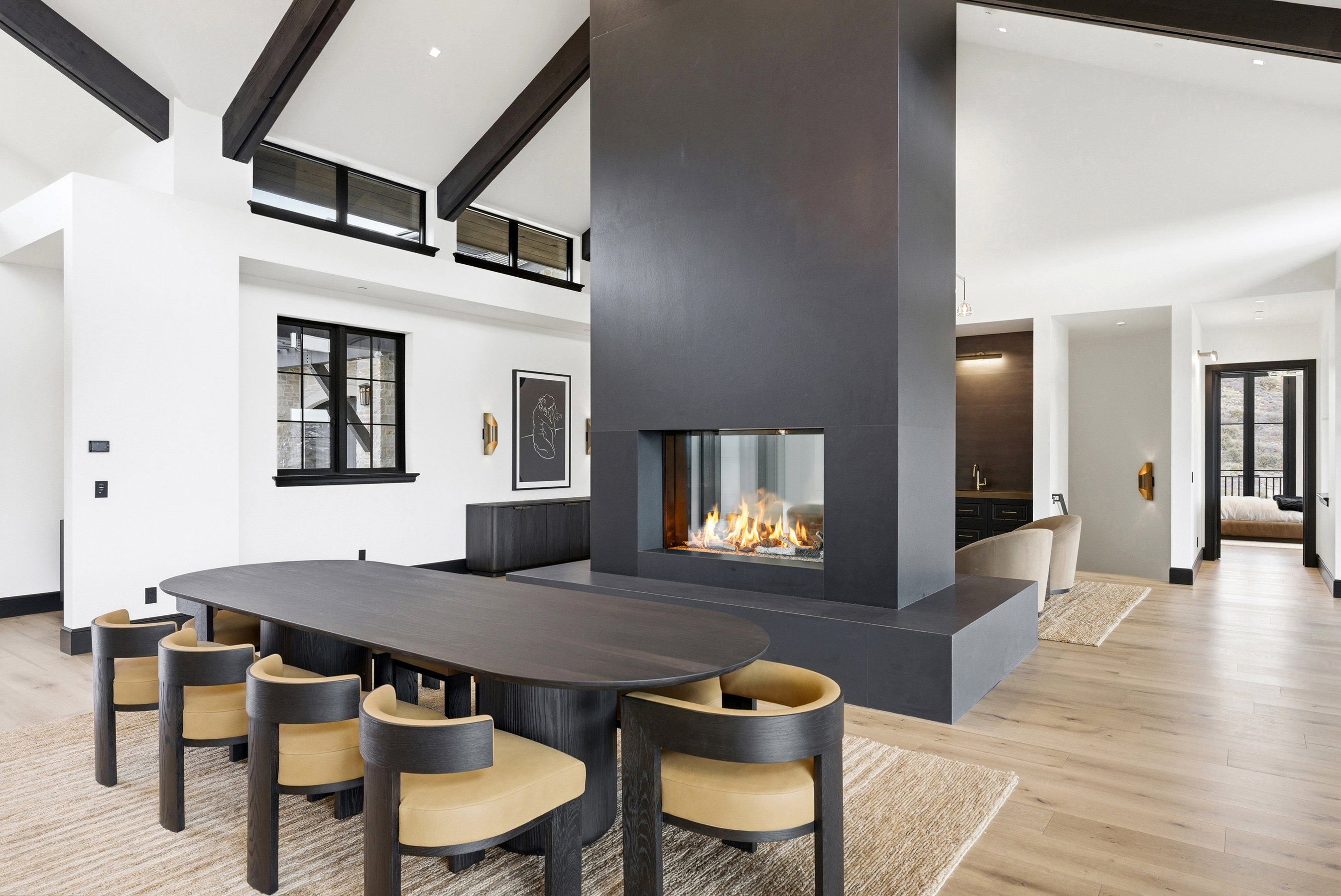 Contemporary open-concept interior featuring a prominent three-sided fireplace with a hearth, vaulted ceilings with exposed dark wood beams, and light wood-finish flooring