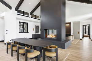 Contemporary open-concept interior featuring a prominent three-sided fireplace with a hearth, vaulted ceilings with exposed dark wood beams, and light wood-finish flooring