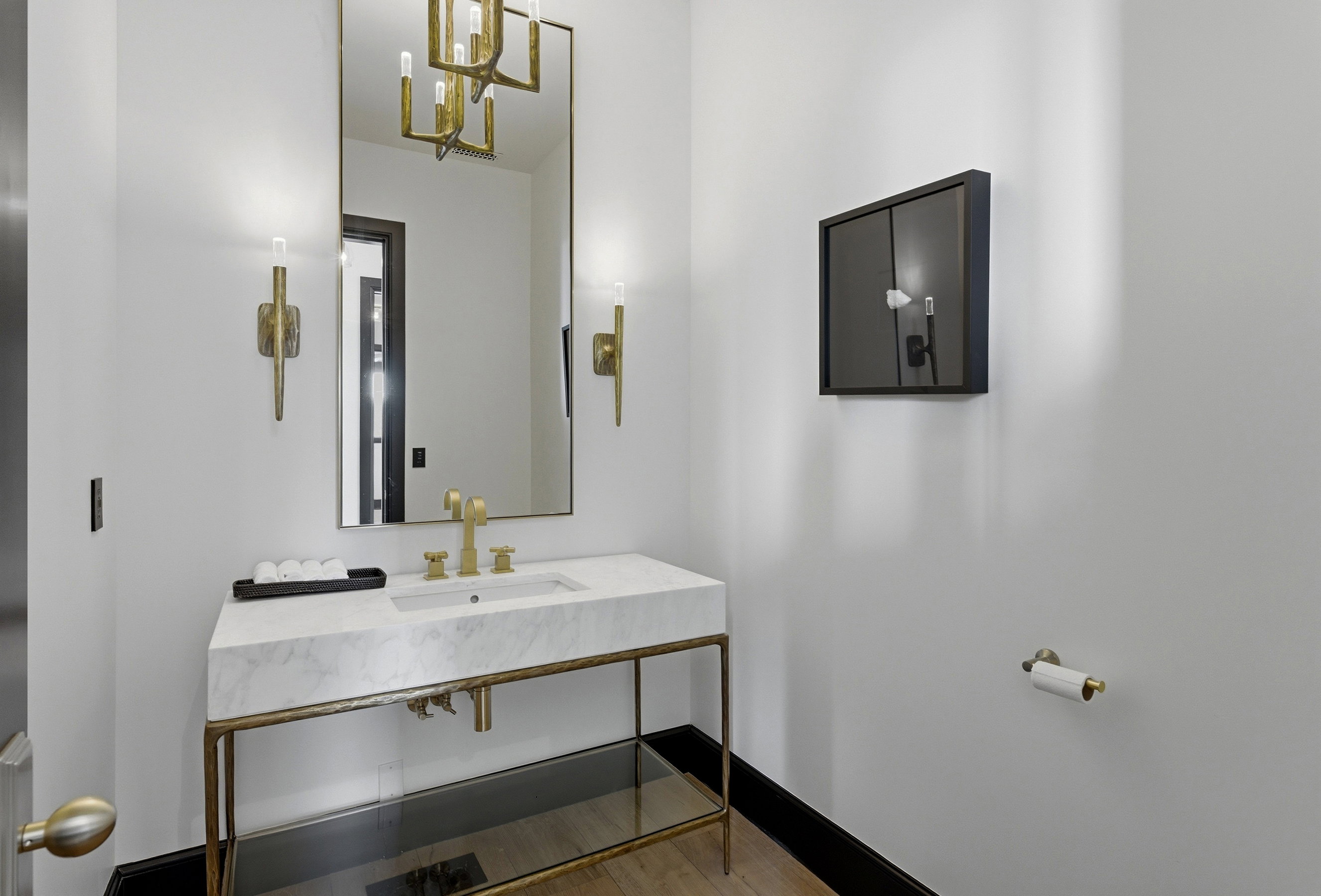 Powder room featuring a rectangular vanity mirror, dual gold-toned wall sconces, a gold-toned chandelier reflection, a white marble countertop vanity with gold-toned fixtures, and a black baseboard
