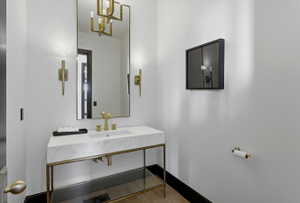Powder room featuring a rectangular vanity mirror, dual gold-toned wall sconces, a gold-toned chandelier reflection, a white marble countertop vanity with gold-toned fixtures, and a black baseboard