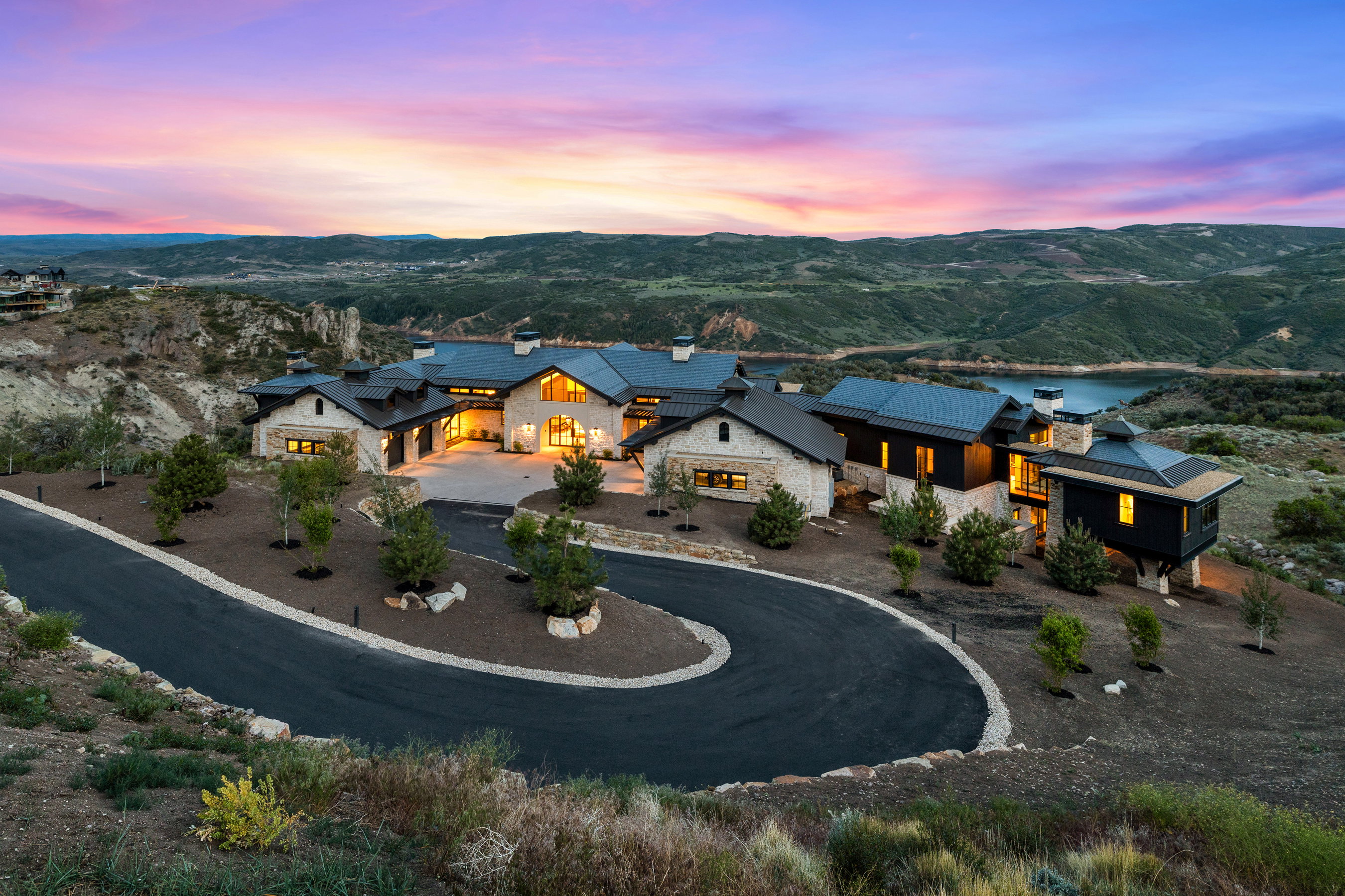 Expansive estate featuring a stone and dark wood exterior, multiple gables, a paved driveway, mature landscaping, and an elevated position overlooking a body of water