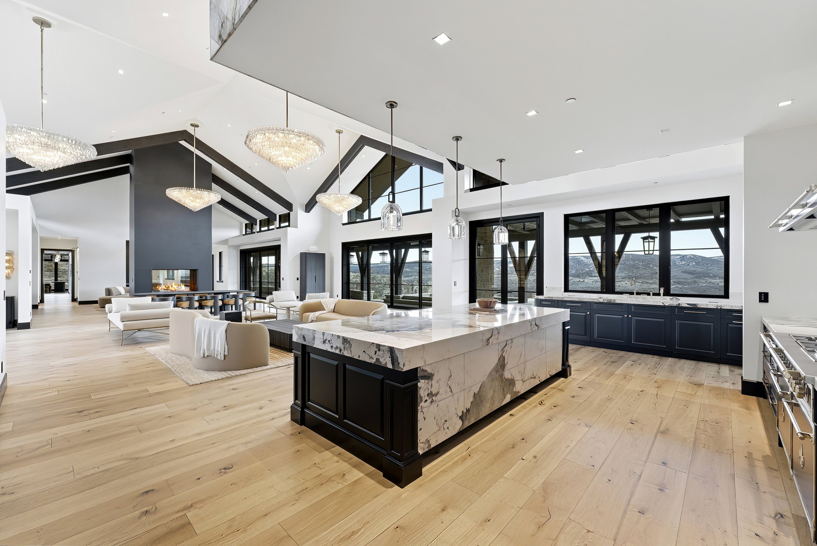 Open-concept living space featuring vaulted ceilings with exposed beams, wood-finish flooring, a contemporary fireplace, and an oversized kitchen island with a stone countertop