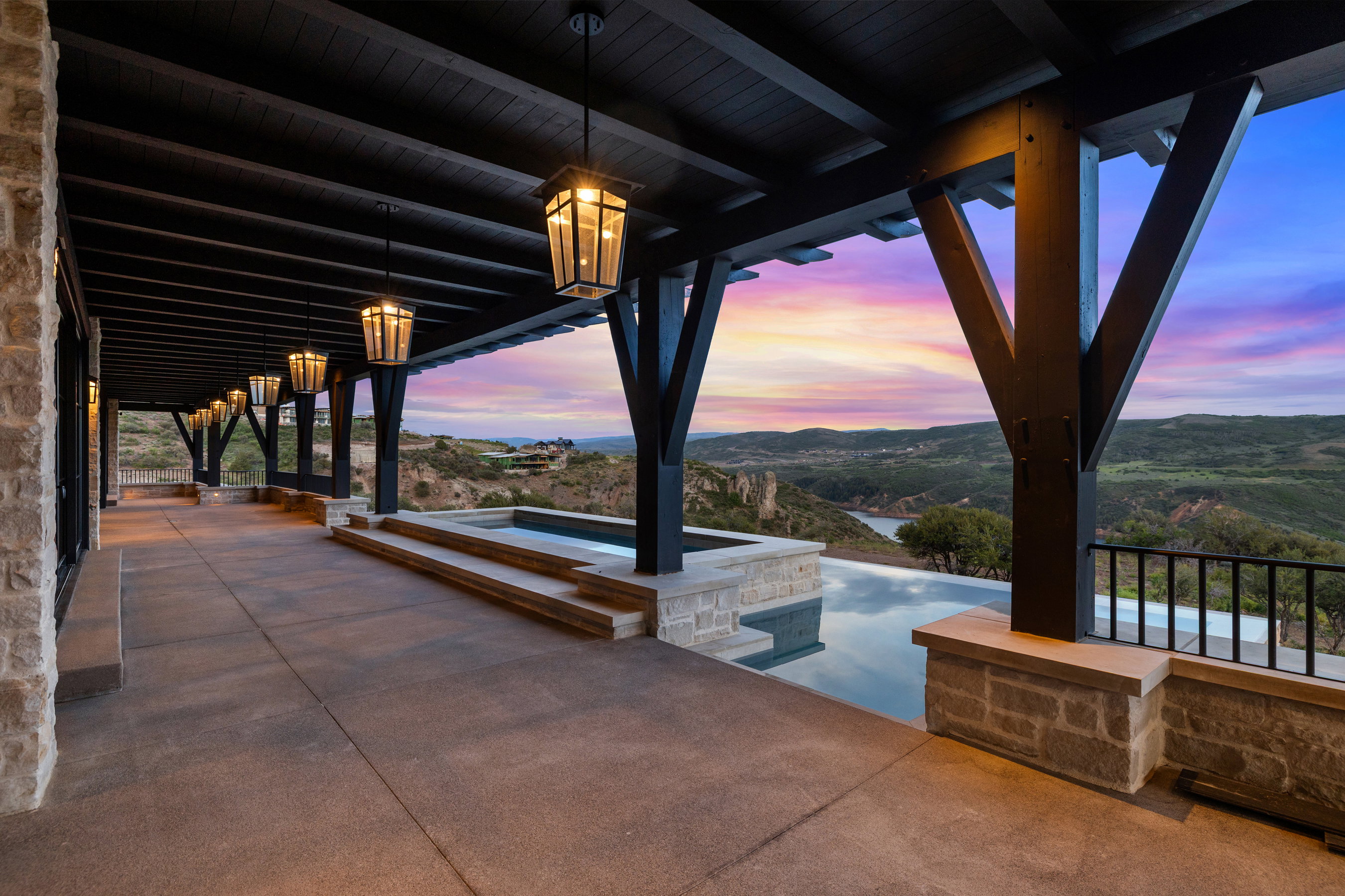 Expansive covered patio featuring a dark wood beam ceiling with pendant lighting, a stone exterior wall, and a multi-level infinity-edge pool
