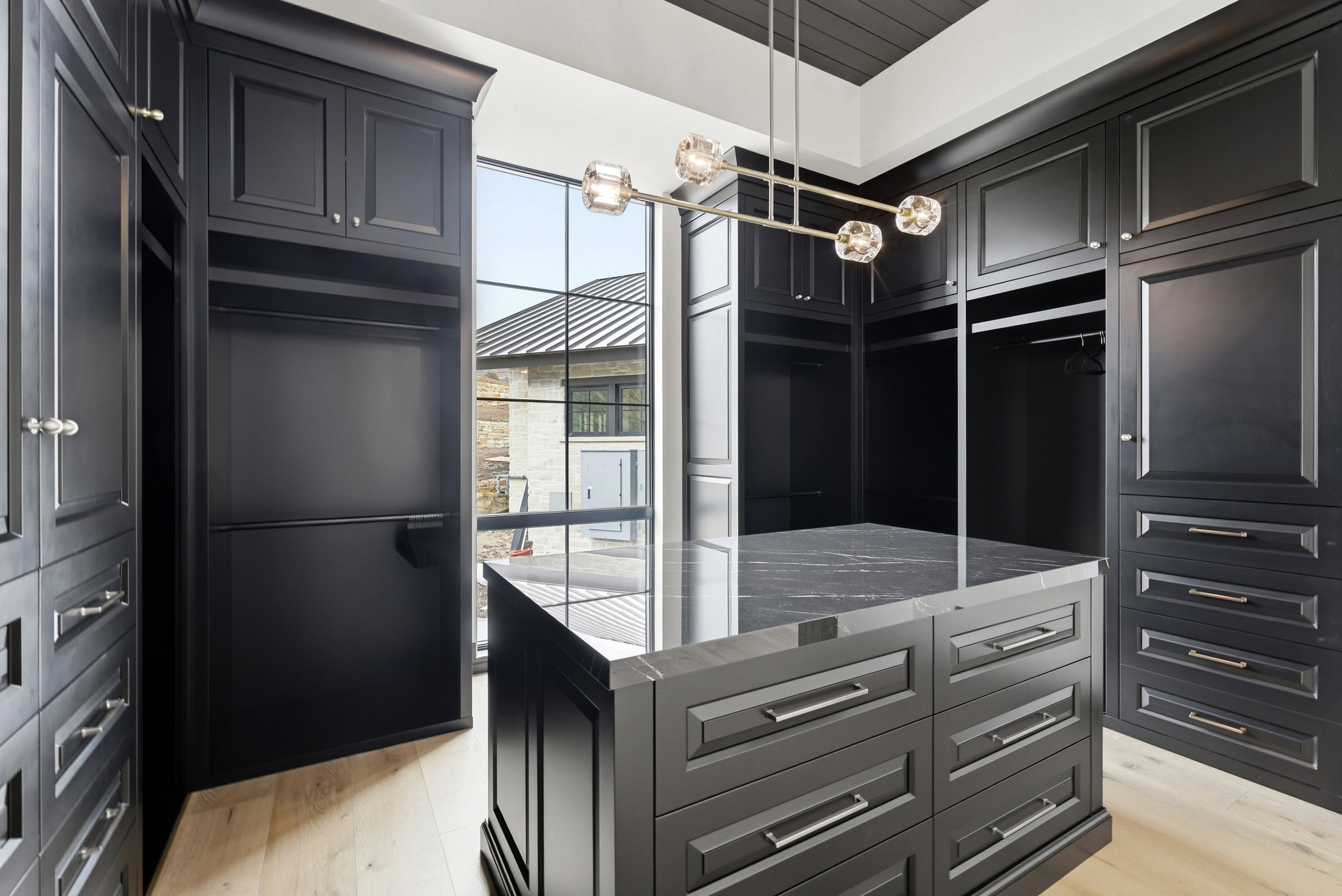 Custom walk-in closet featuring extensive built-in cabinetry, a central island with a stone countertop, a contemporary chandelier, large picture windows, and light wood-finish flooring