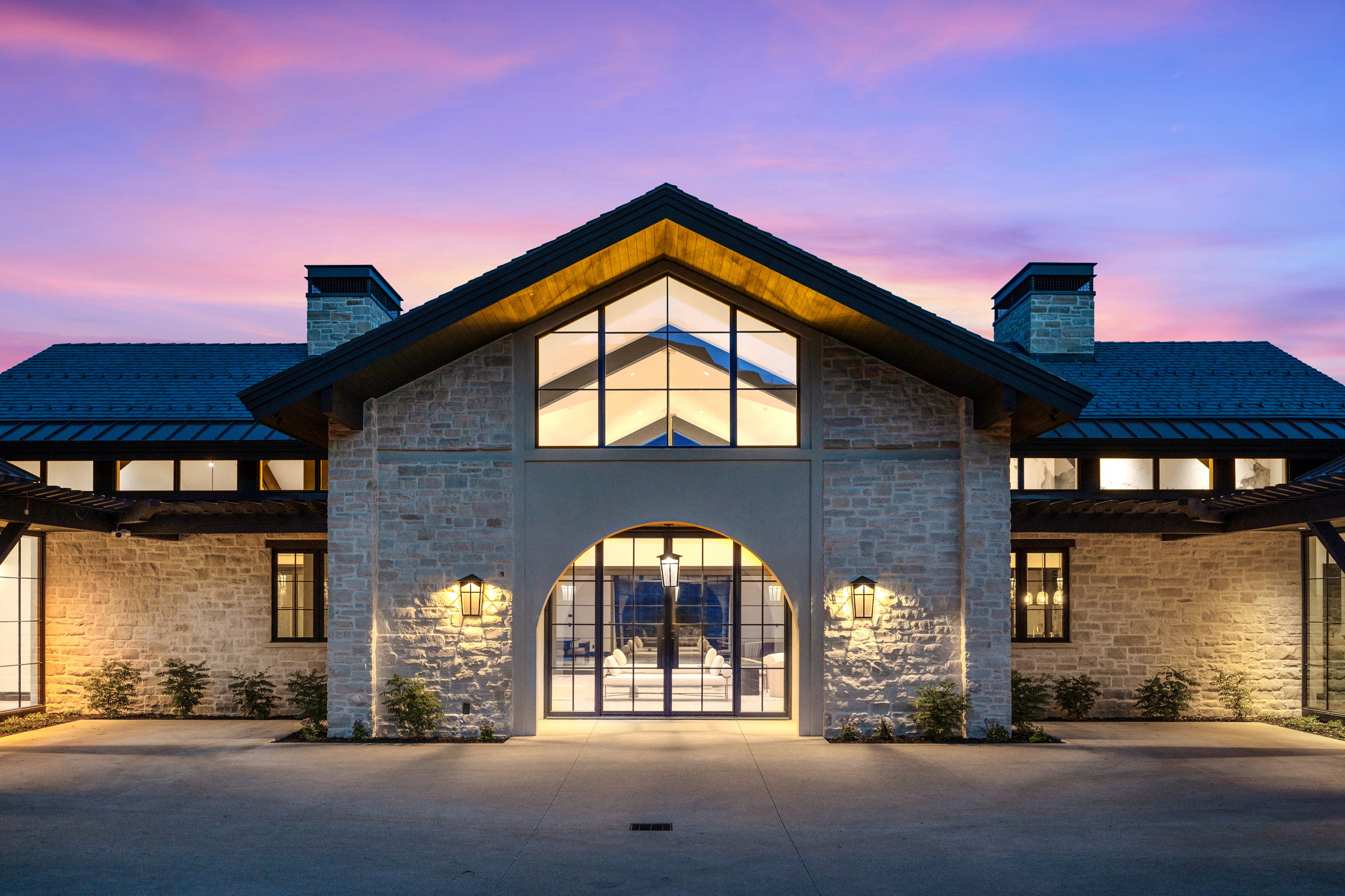 Stone facade residence featuring a prominent arched entrance with glass paneled double doors, a gabled roofline, and an expansive concrete driveway