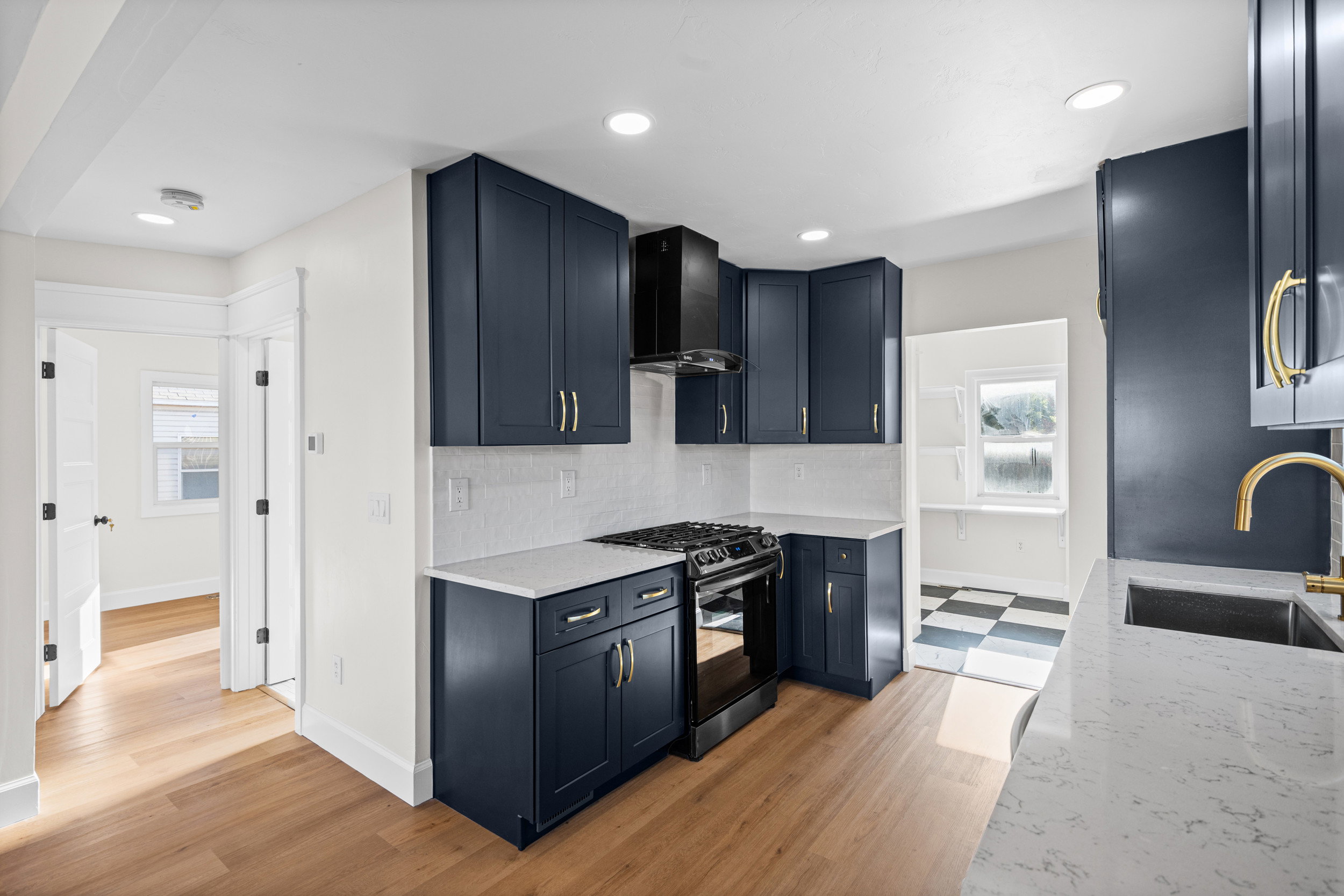 Kitchen featuring dark blue cabinetry, light-colored countertops, wood-finish flooring, a stainless steel sink with a gold-finish faucet, and a black gas range with matching hood