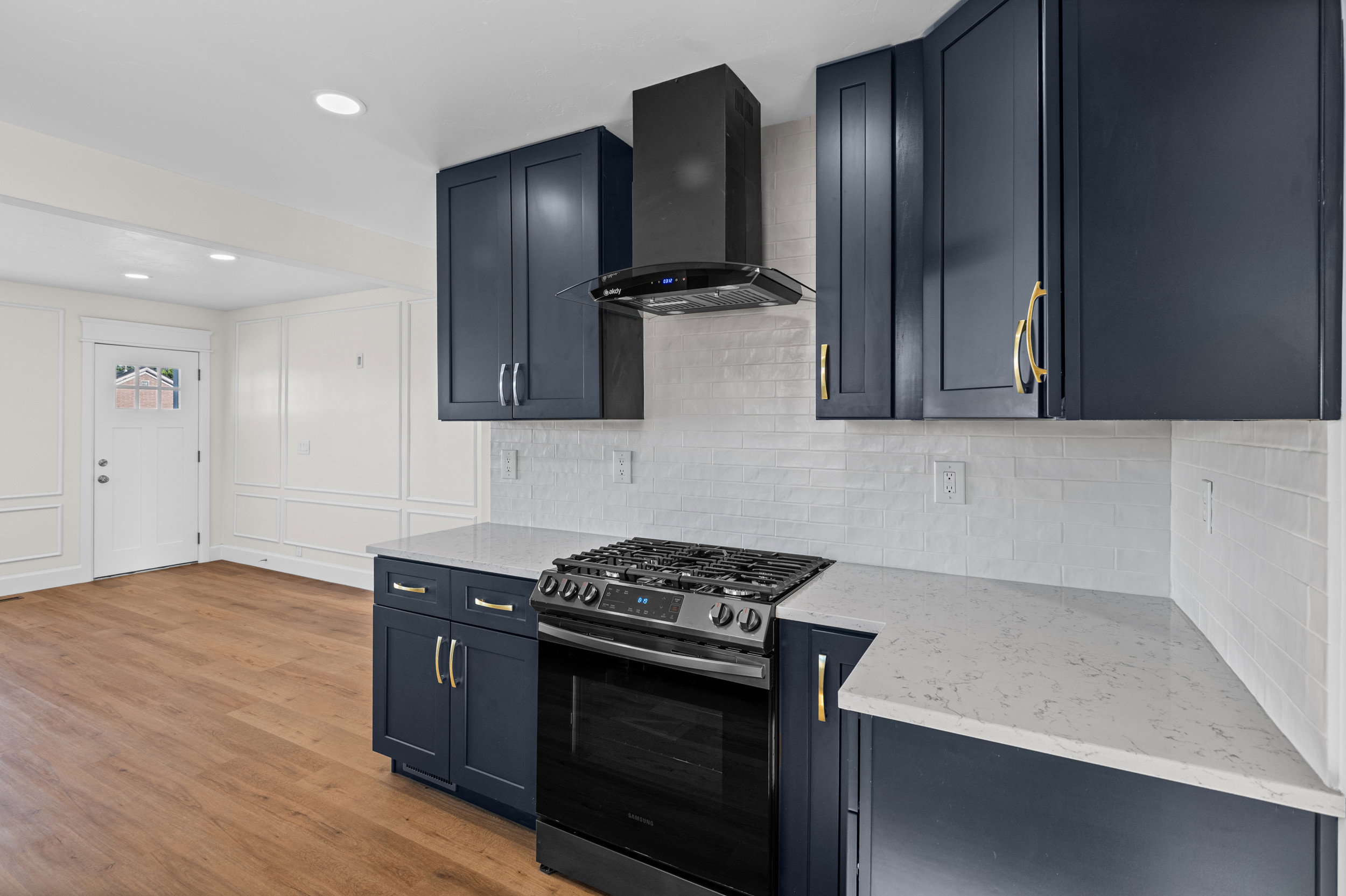 Kitchen featuring dark blue cabinetry with gold-tone hardware, white subway tile backsplash, light-toned countertops, a black range, and a black range hood