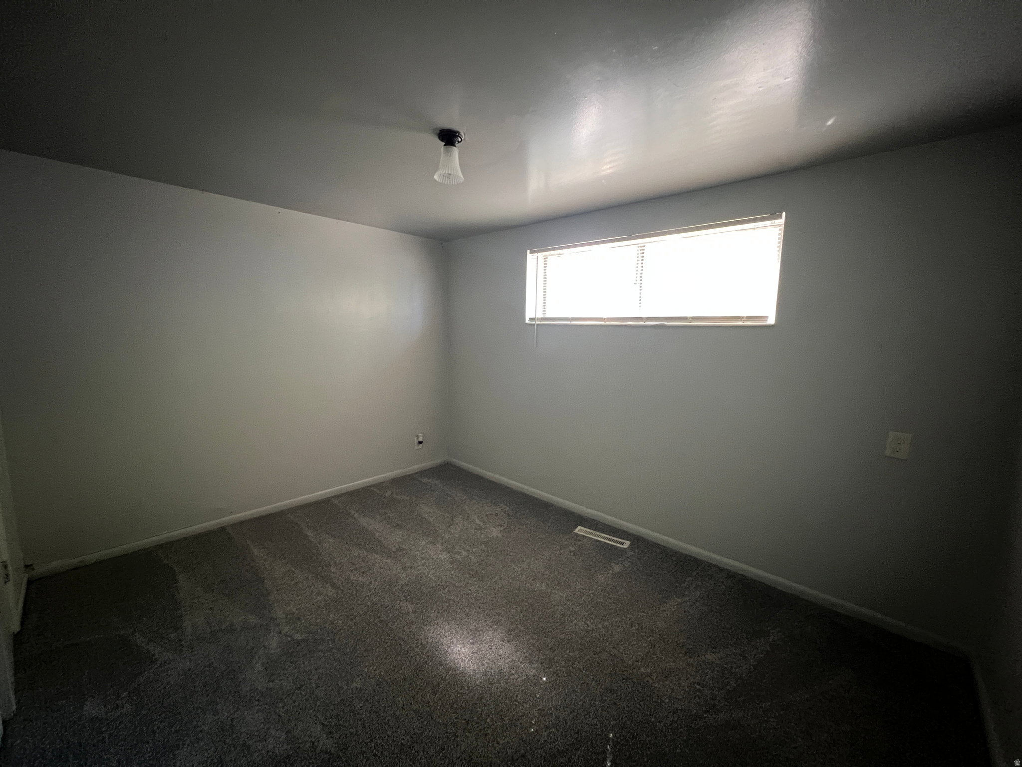 Room featuring neutral gray wall paint, dark gray patterned carpeting, a single horizontal window with blinds, and a ceiling-mounted light fixture