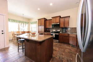 Kitchen featuring wood cabinetry, stainless steel appliances, a patterned tile backsplash, and a central island with a stone-finish countertop