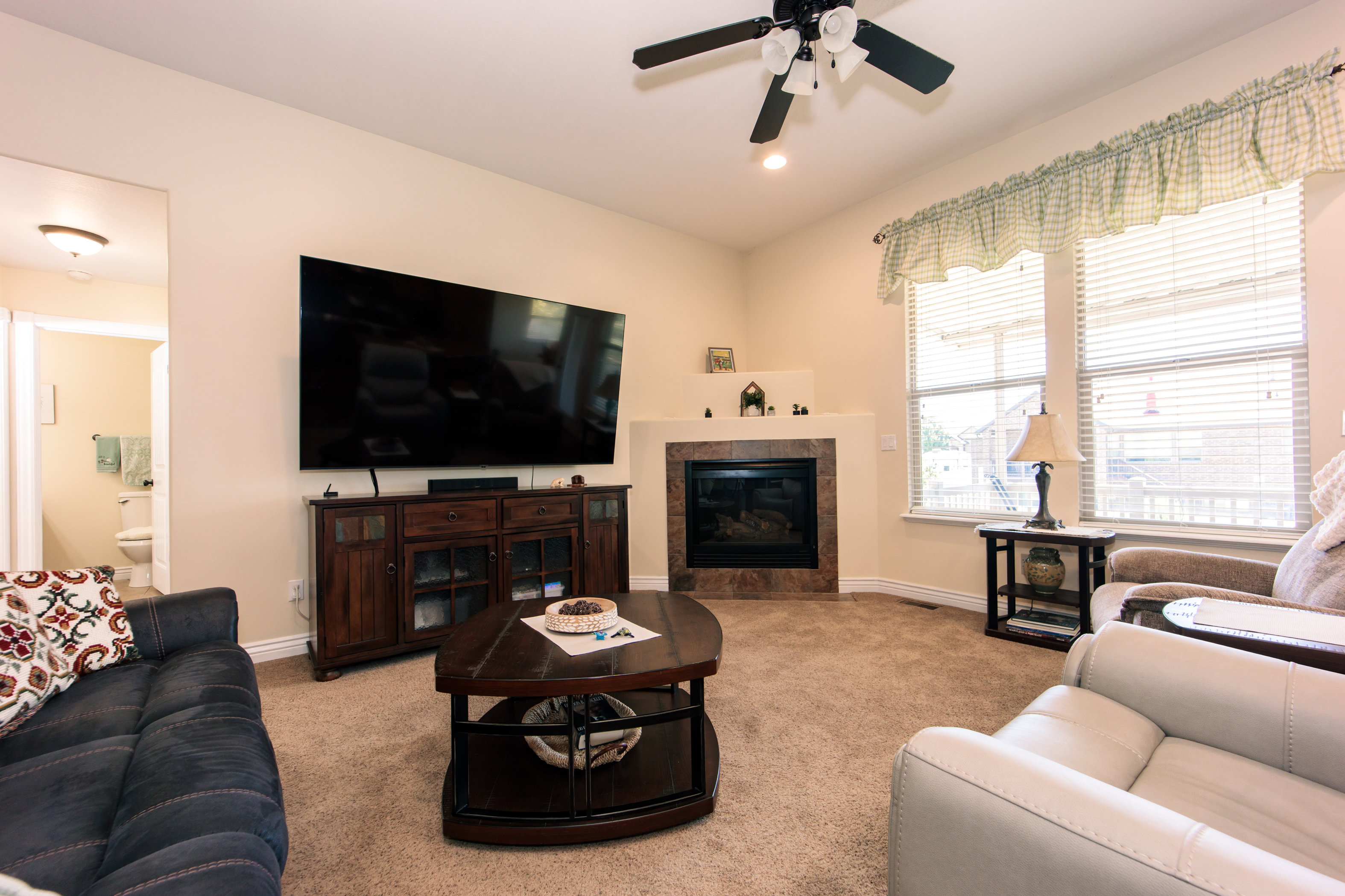 Living room featuring a corner fireplace with a dark stone surround and hearth, carpeted flooring, and a ceiling fan with integrated lighting