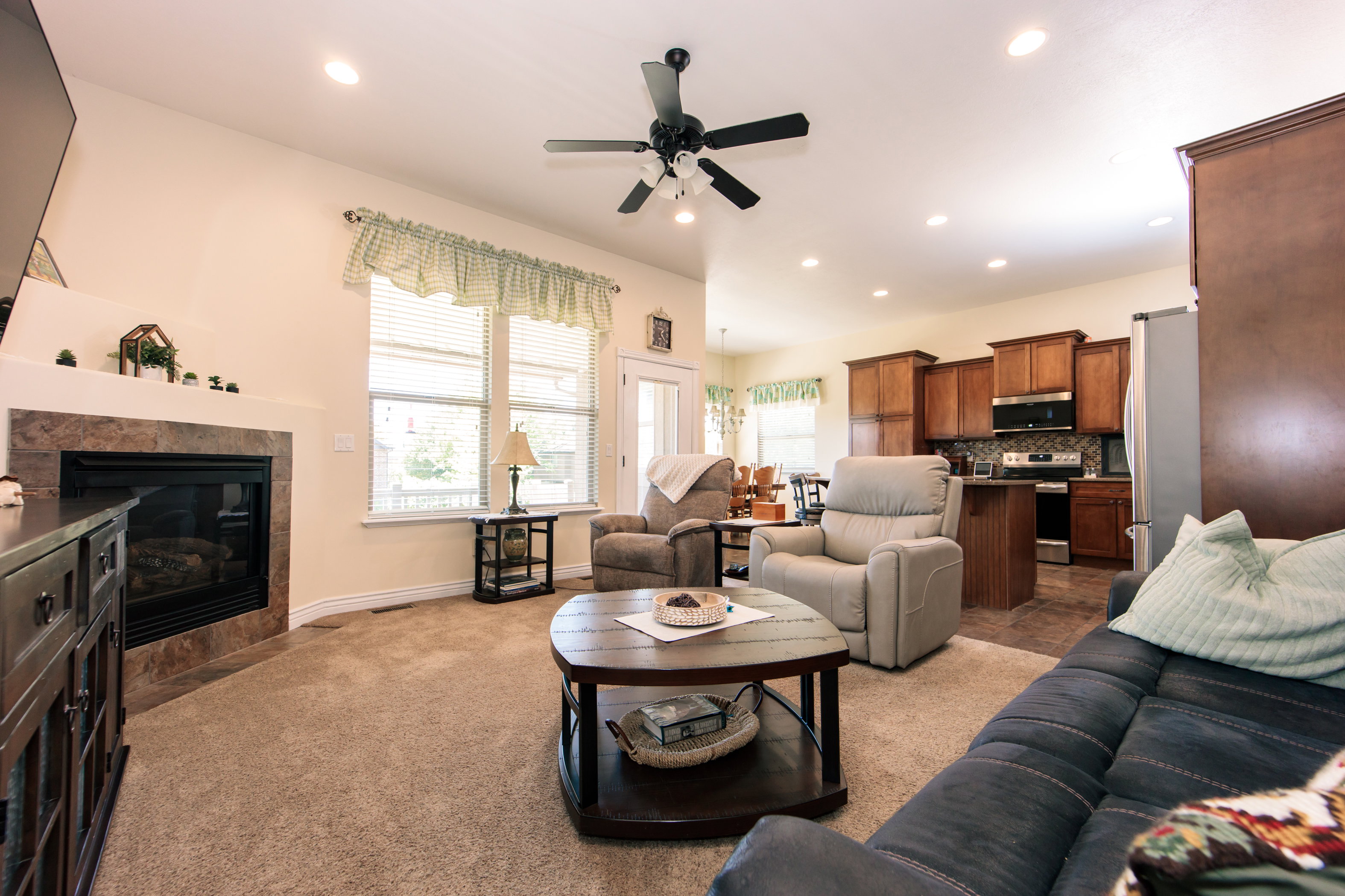 Open-concept living space featuring a gas fireplace with a stone-tile surround, neutral carpeting, and a ceiling fan with integrated lighting