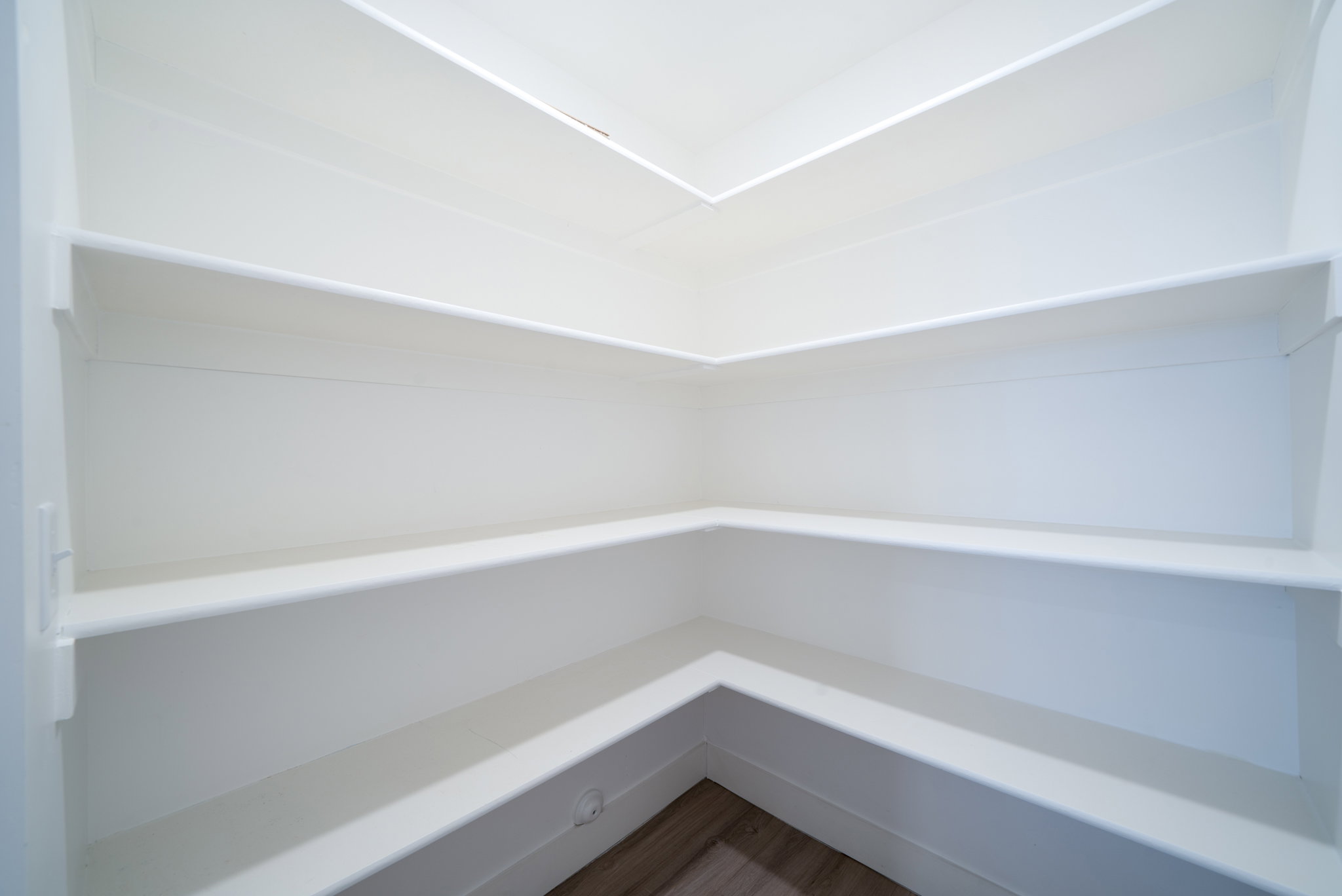 Spacious corner pantry featuring multiple tiers of white shelving and wood-finish flooring