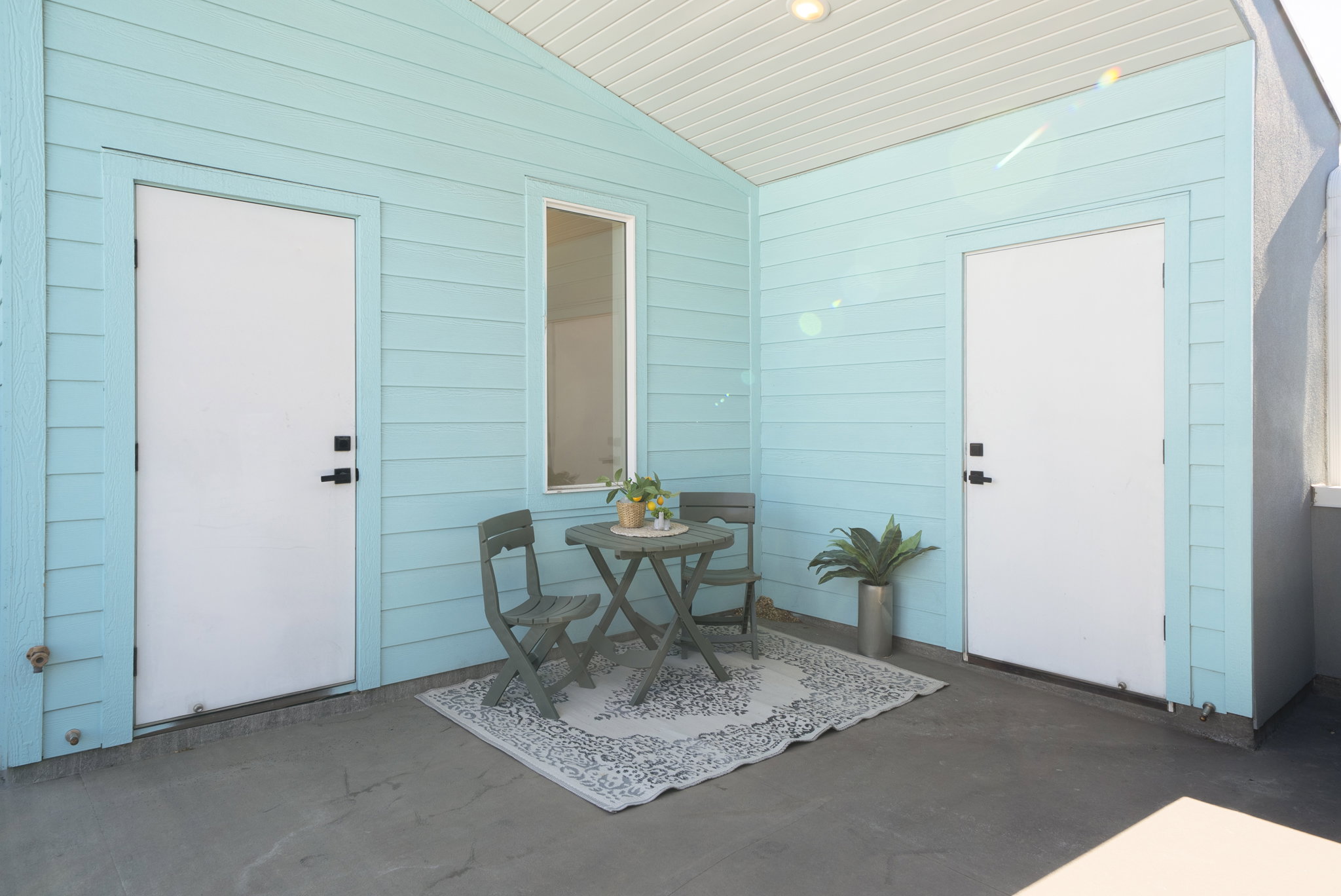 Covered outdoor patio area featuring light blue siding, a single fixed window, and two white entry doors with matte black hardware