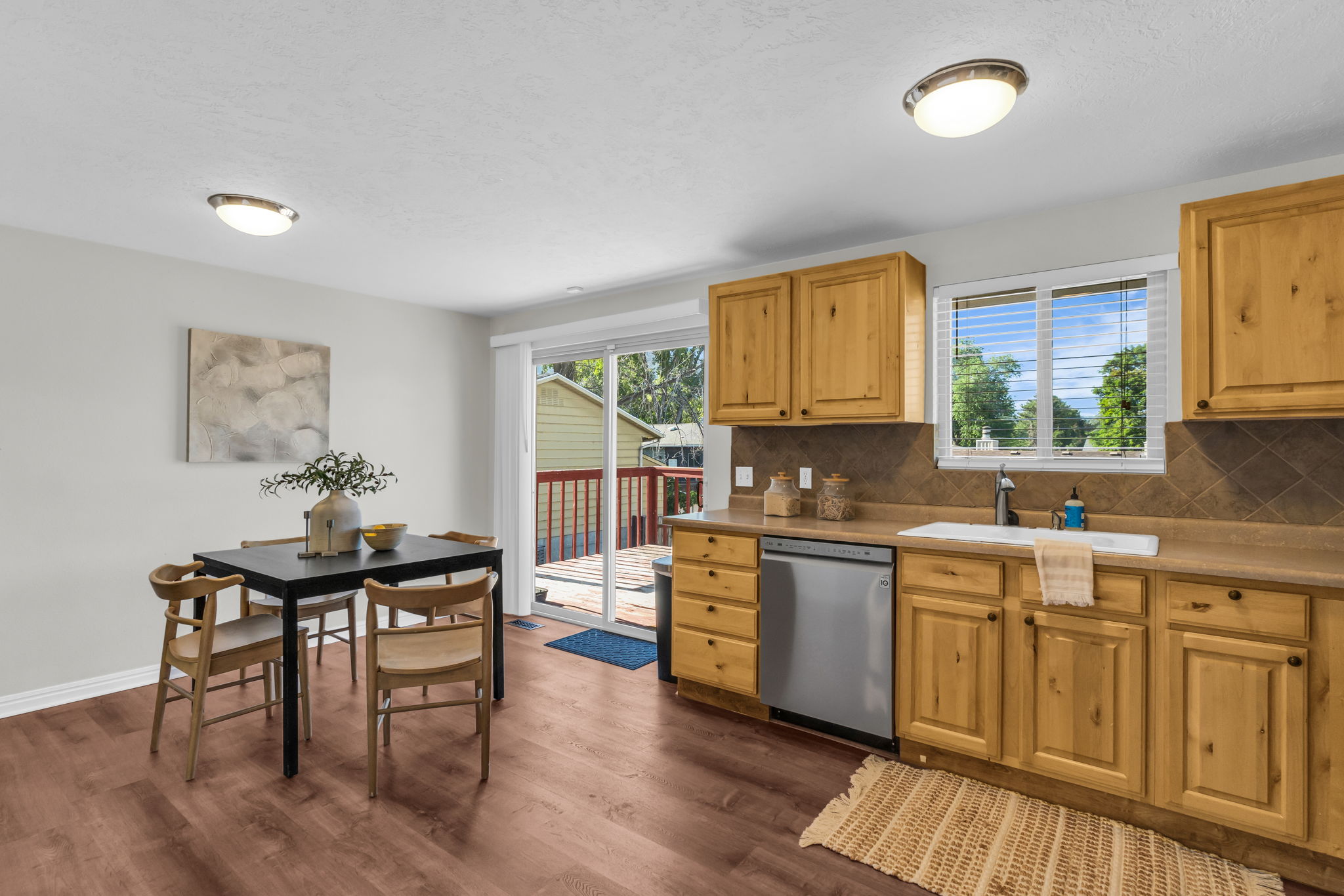Kitchen and dining area featuring wood-finish flooring, light wood cabinetry, a tile backsplash, and a stainless steel dishwasher