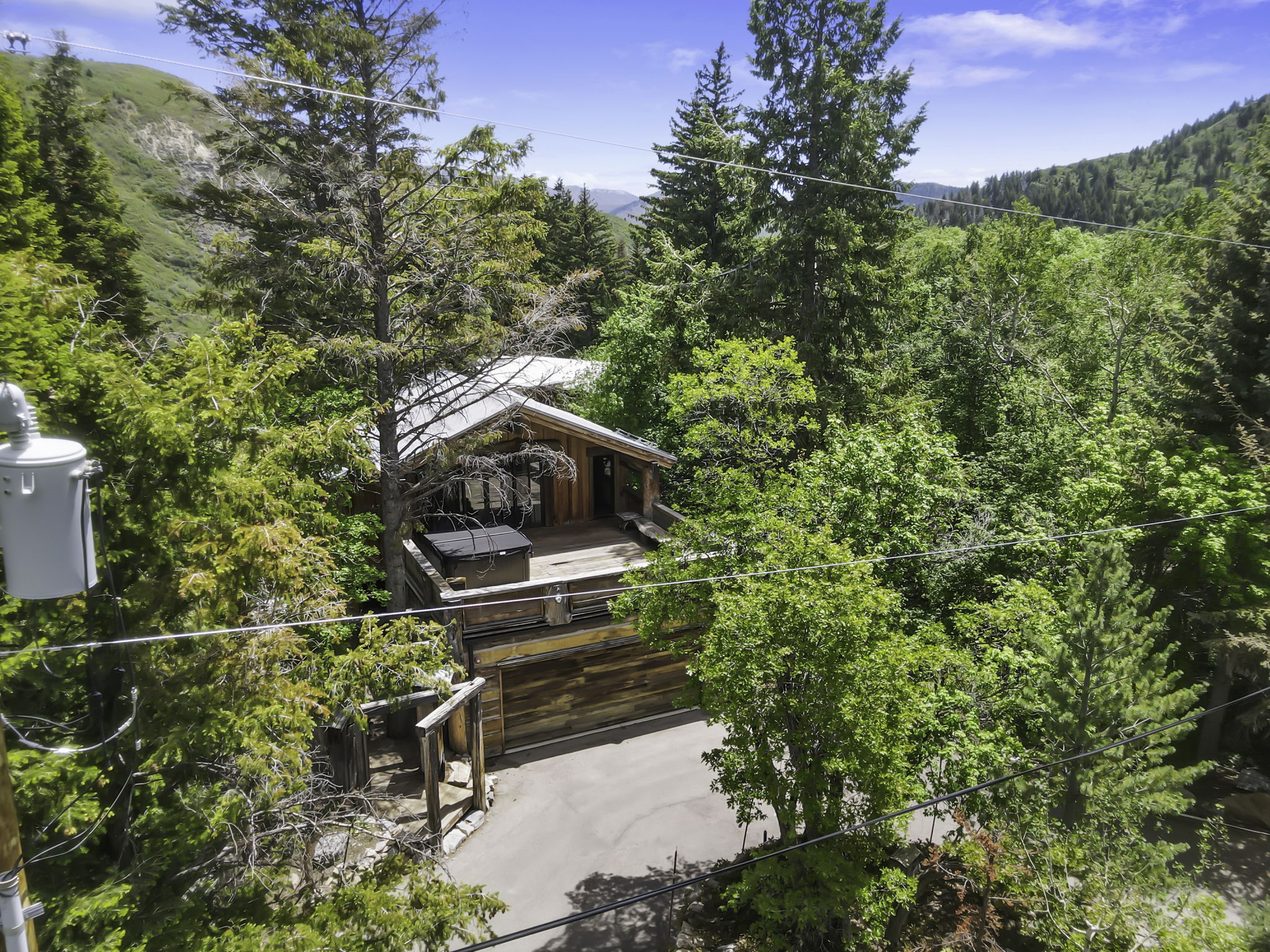 Wood-sided cabin exterior with a large wooden deck and an integrated hot tub, surrounded by mature trees and a paved driveway