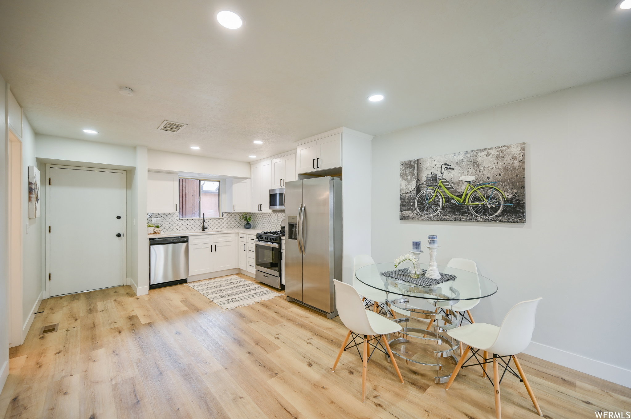 Open-concept kitchen and dining area featuring light wood-finish flooring, white cabinetry, stainless steel appliances, a geometric tile backsplash, and recessed lighting