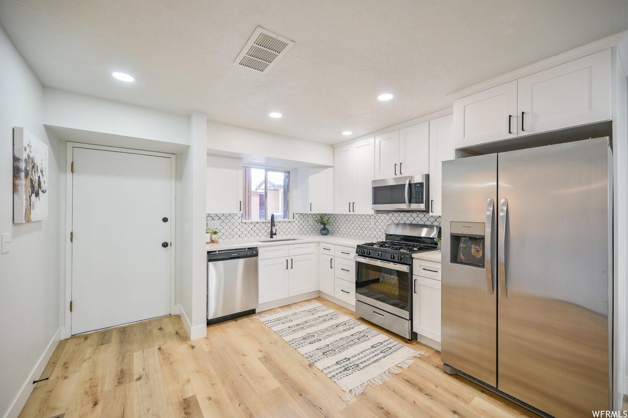 Modern kitchen featuring white shaker cabinetry with black hardware, stainless steel appliances, a geometric tile backsplash, and light wood-finish flooring
