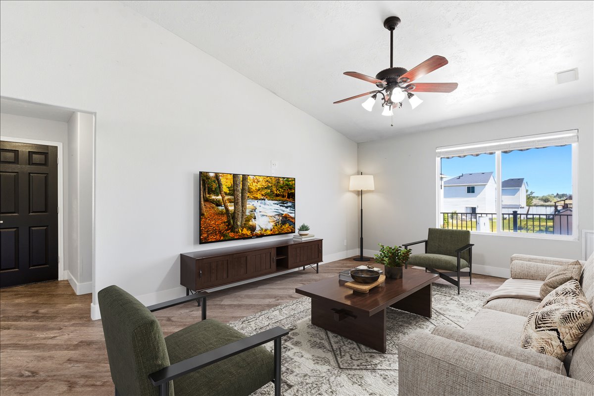 Living area featuring a vaulted ceiling with a ceiling fan, a large window providing natural light, wood-finish flooring, and a six-panel door with matte black hardware Virtually staged