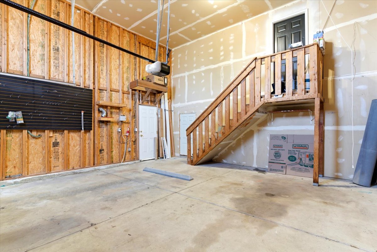 Spacious garage featuring a finished concrete floor, exposed wood framing, an automatic garage door opener, and a wooden staircase leading to an upper landing with a dark panel door