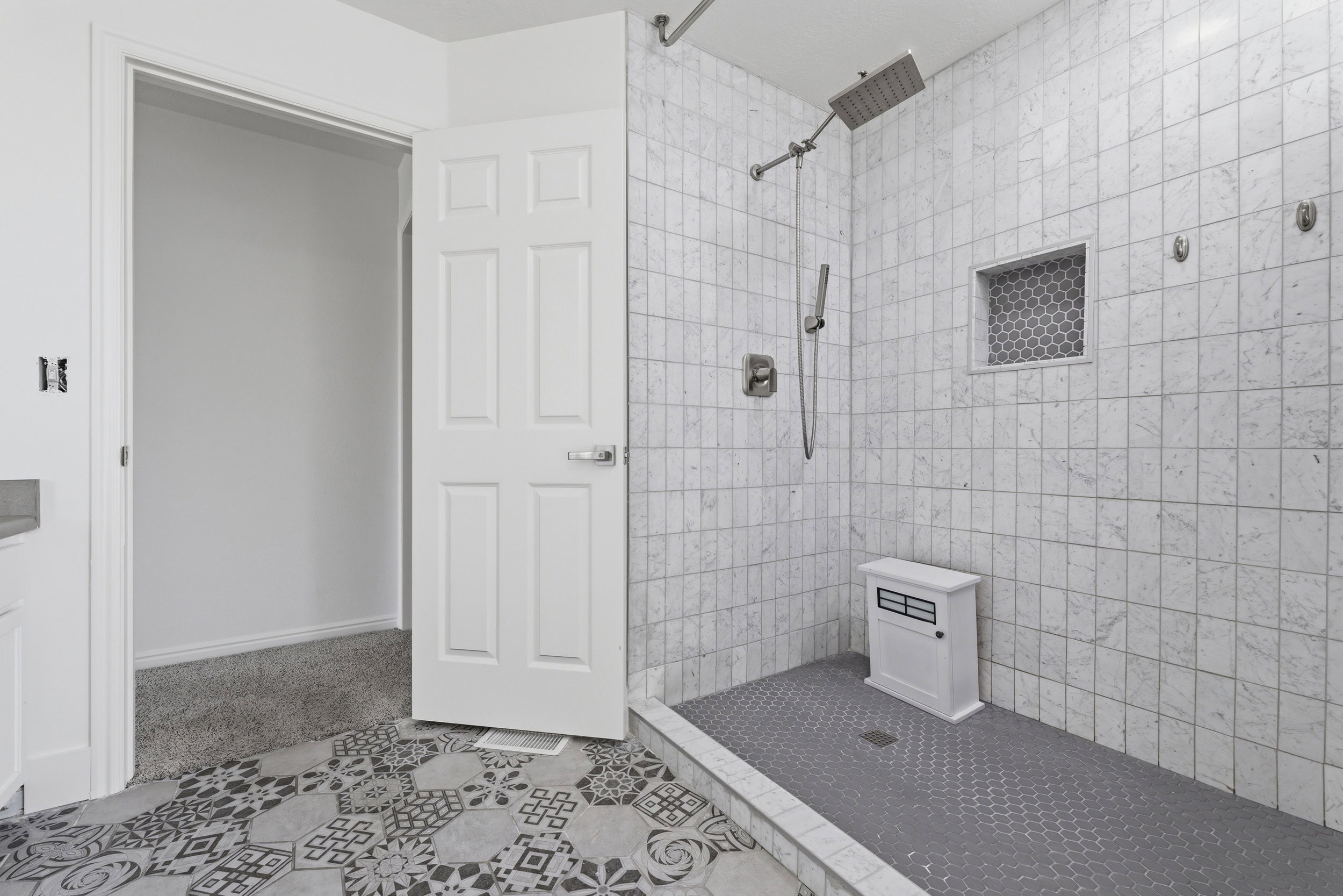 Walk-in shower featuring gray hexagonal floor tiling, rectangular wall tiling, a built-in niche with hexagonal accent tiles, and a rainfall showerhead with a handheld sprayer