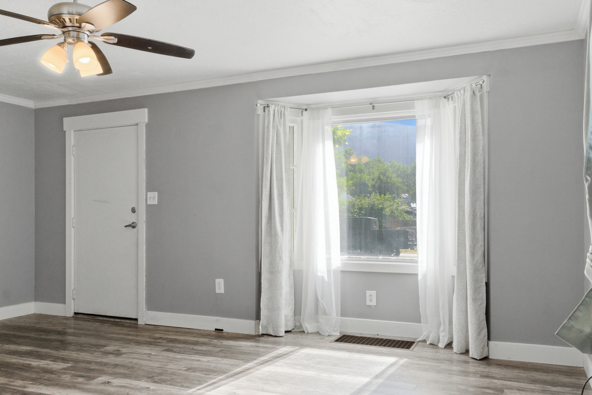 Bright living area featuring large front window, gray walls, white trim, ceiling fan, and LVP flooring.