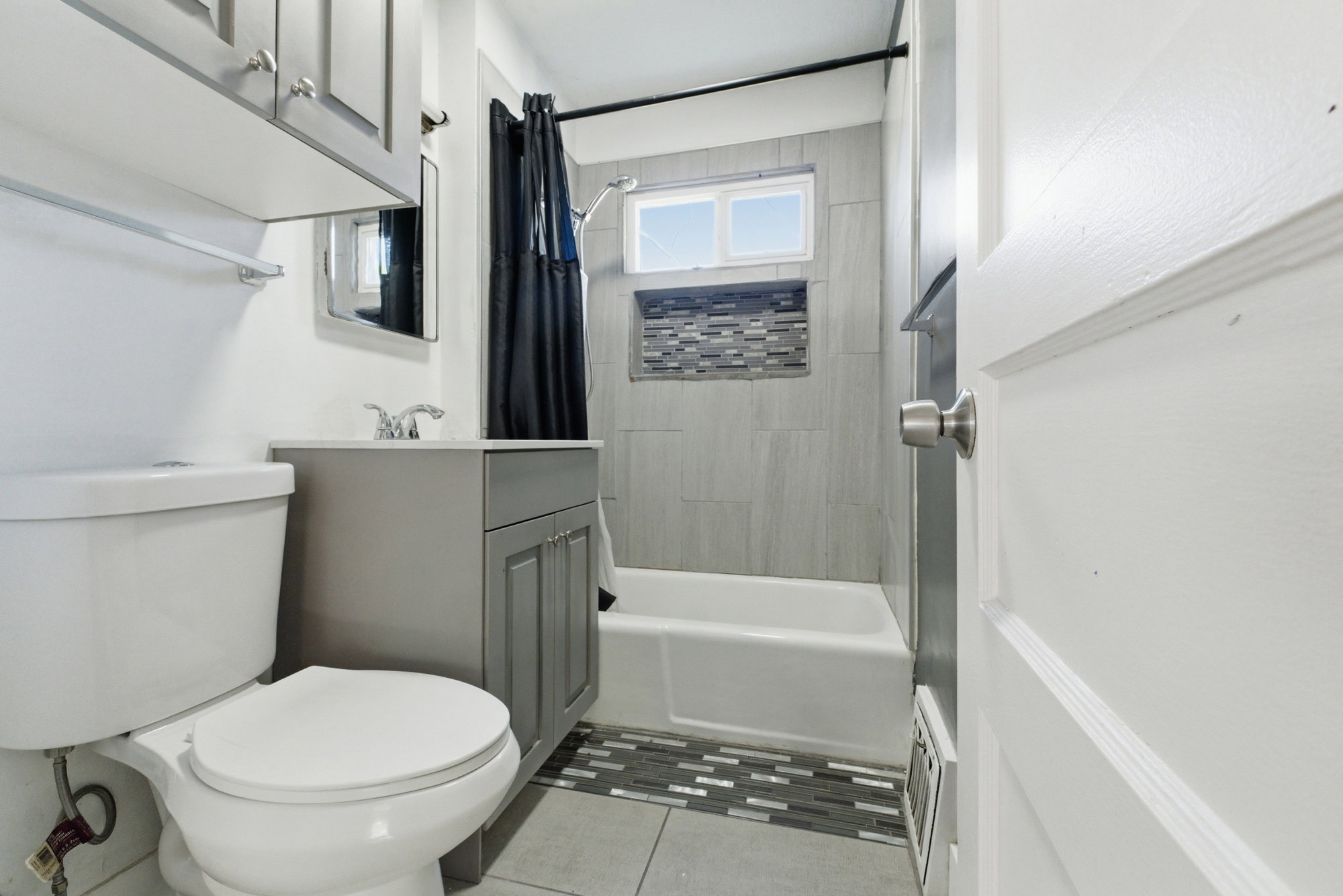 Updated bathroom with tub/shower combo, tile surround, decorative accent niche, gray vanity, and tile flooring.