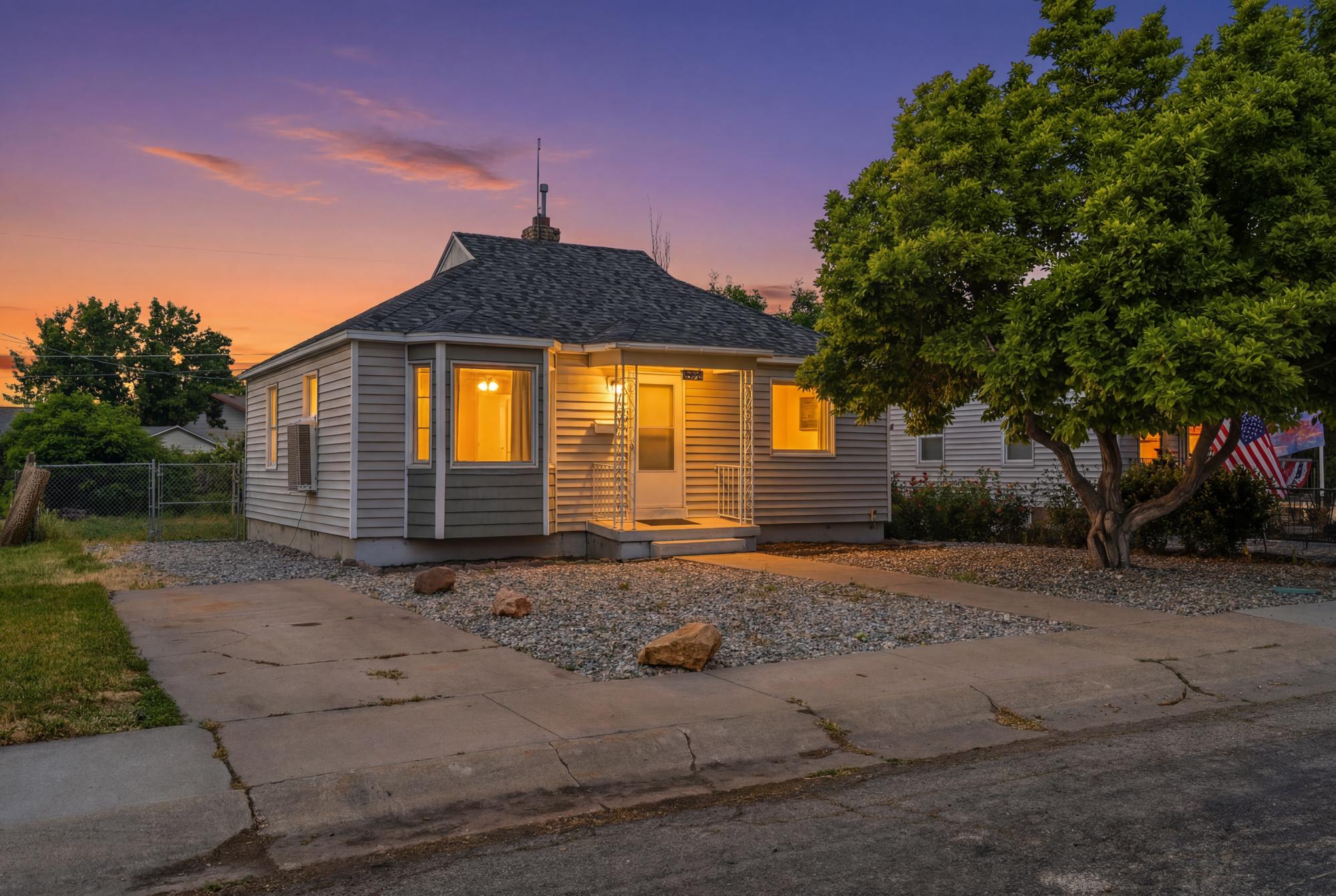 Twilight exterior view showcasing charming curb appeal, welcoming covered entry, mature shade tree, driveway parking, and low-maintenance landscaping.