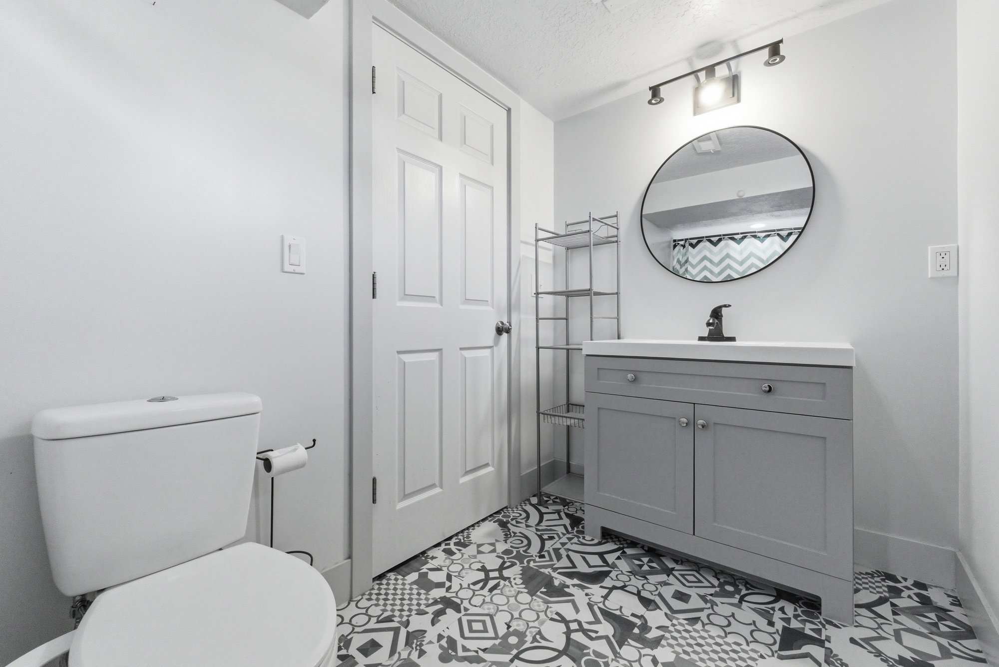 Bathroom vanity area featuring gray cabinet, round mirror, updated light fixture, patterned tile flooring, and storage space.