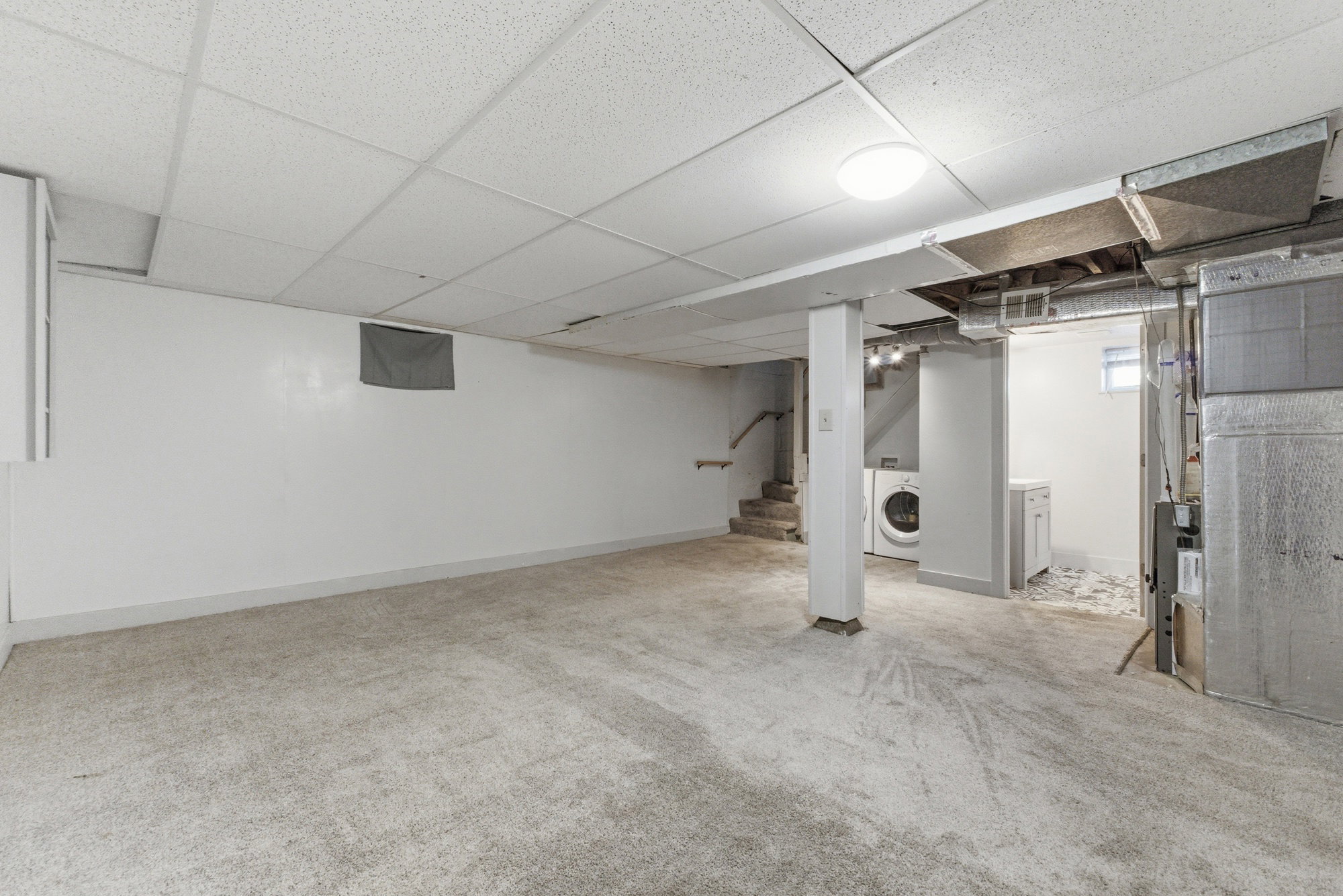 Additional basement view showcasing open finished space, carpet, updated lighting, and access to laundry and utility areas.