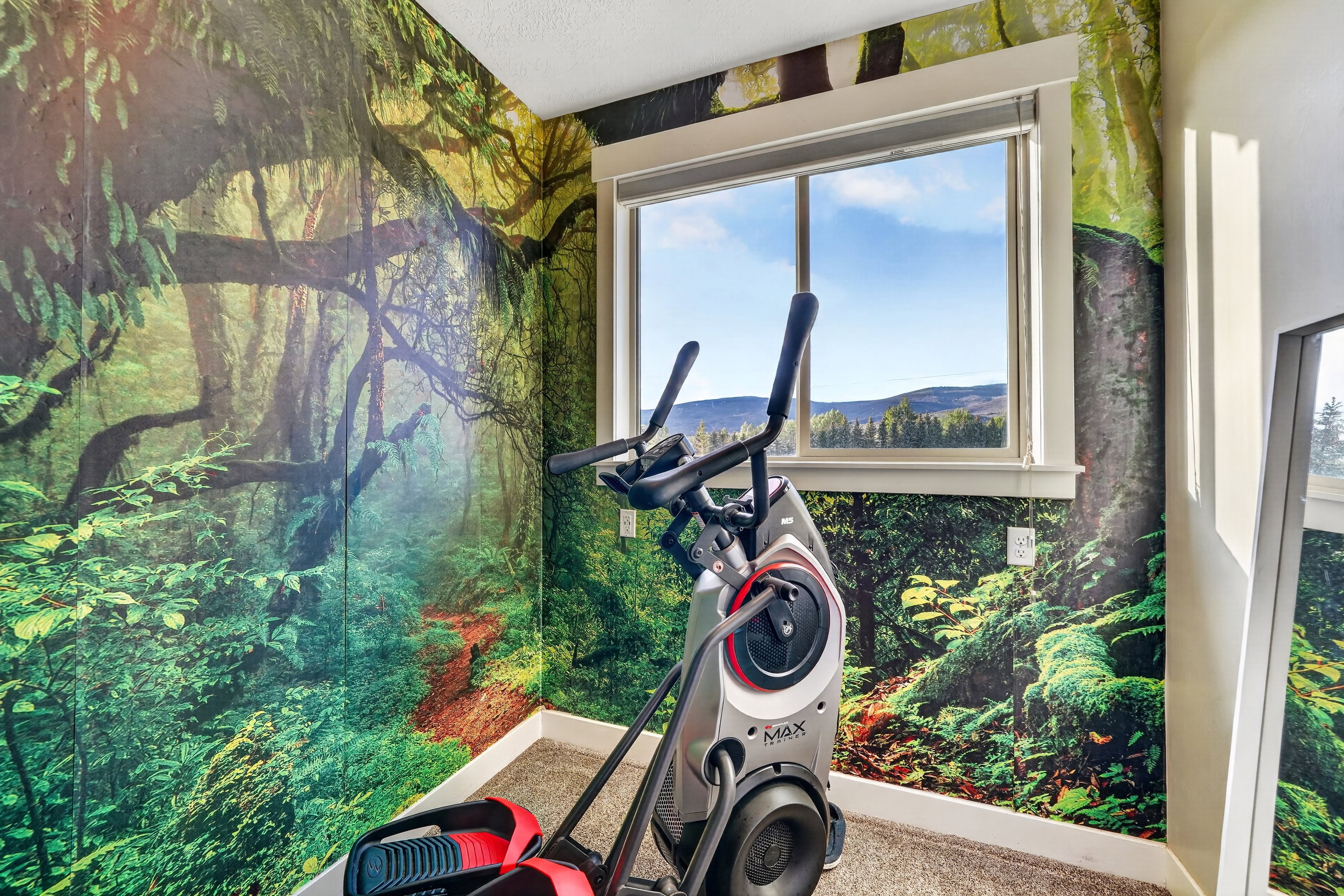 Room featuring vibrant forest-themed wall coverings, a large window, white window trim, and a mountain and sky backdrop