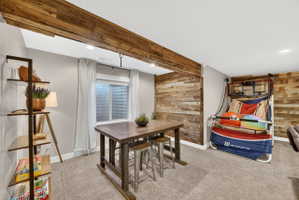 Finished room featuring a wood-finish accent wall, exposed wood-finish beam, recessed lighting, window with privacy screen, and carpeted flooring